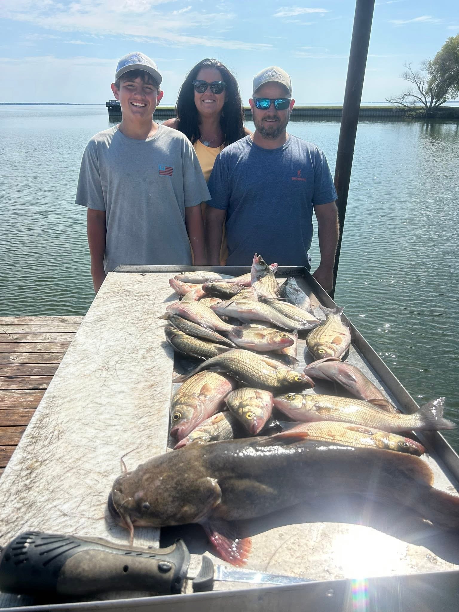 Three people standing behind a table with caught fish on it, near a body of water with a dock and trees in the background.