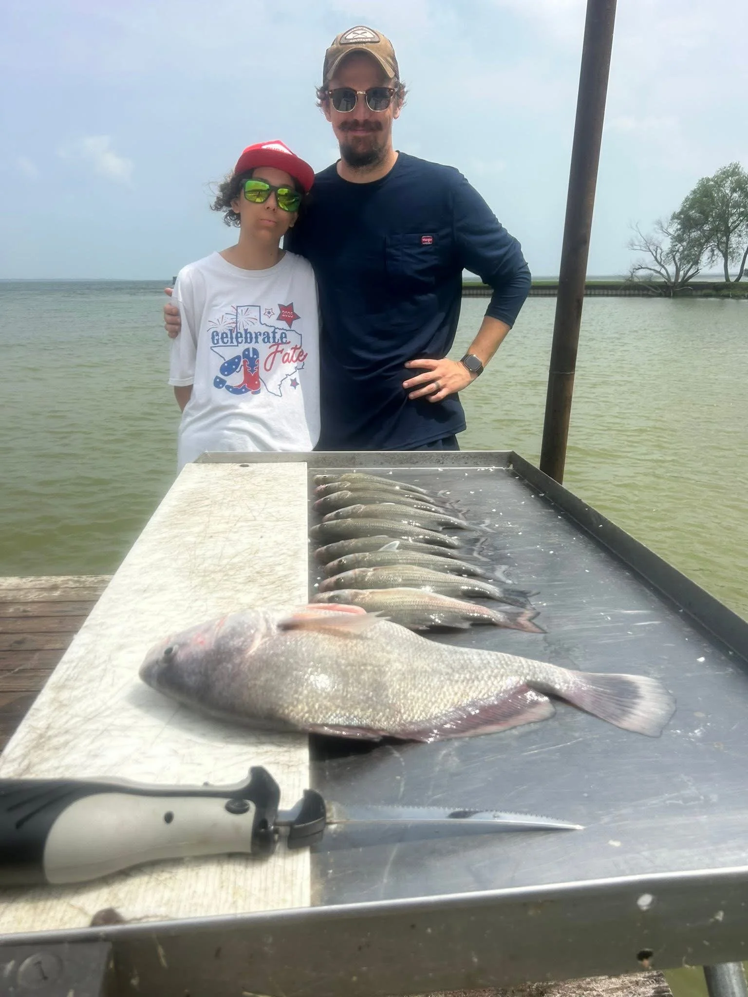 Two people standing outdoors at a water body, with fish laid out on a table in front of them and a knife, possibly after fishing or fishing competition.