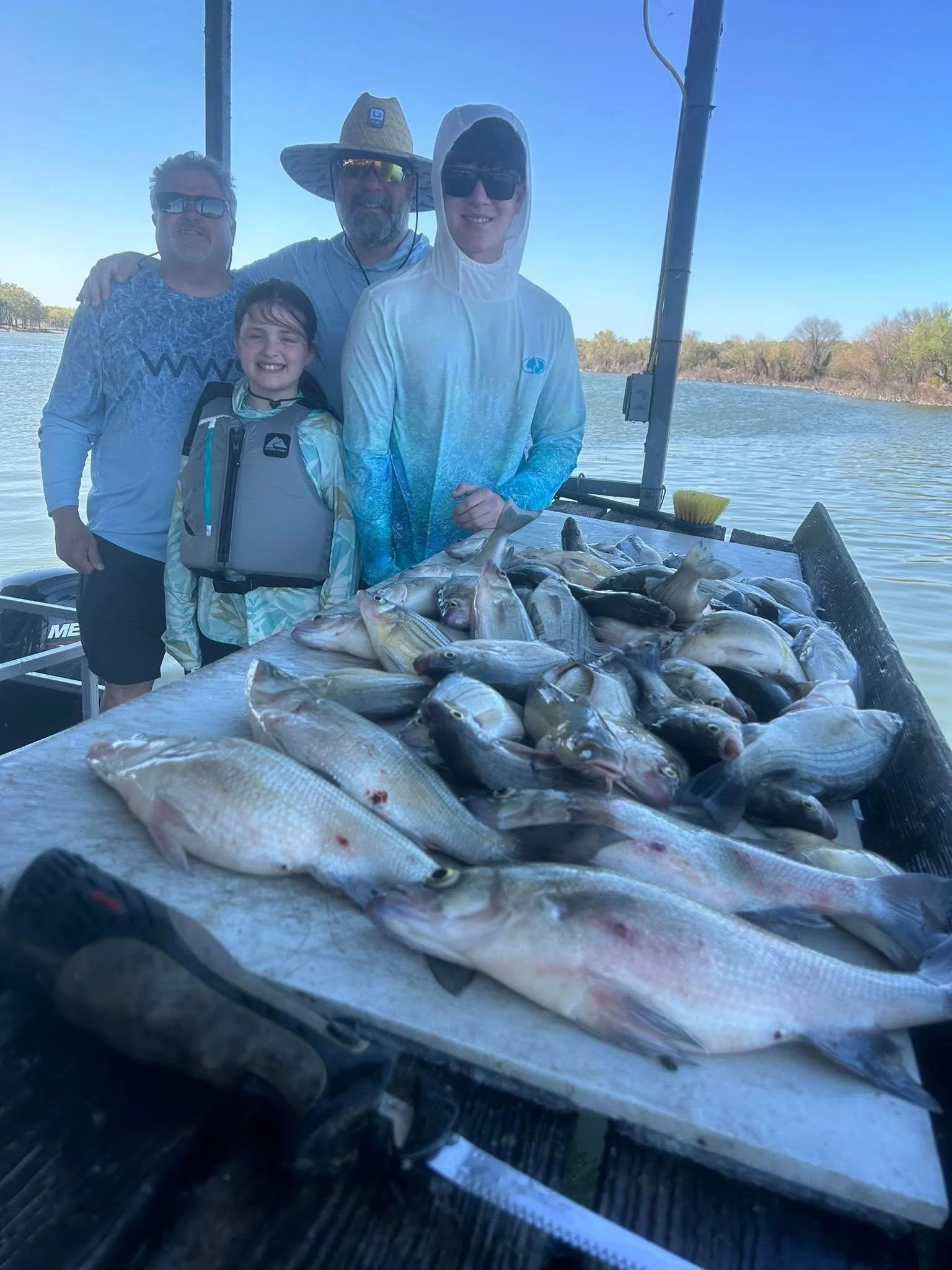 A group of four people on a boat by a river with trees in the background, showing their catch of various fish laid out on a table.