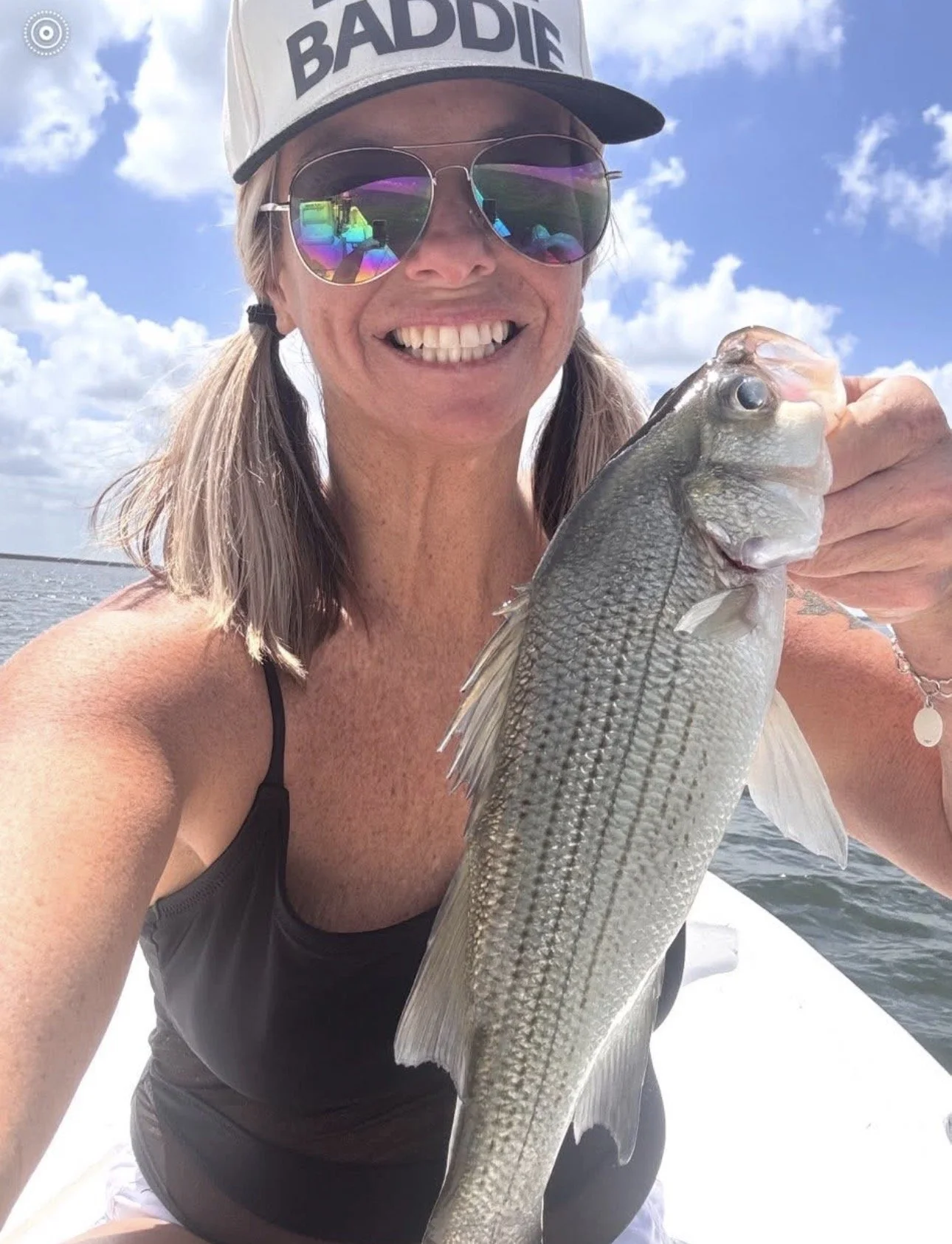 Woman taking a selfie while holding a large caught fish on a boat with water and sky in the background. She is wearing sunglasses, a cap with the word 'BADDIE', and a black top.