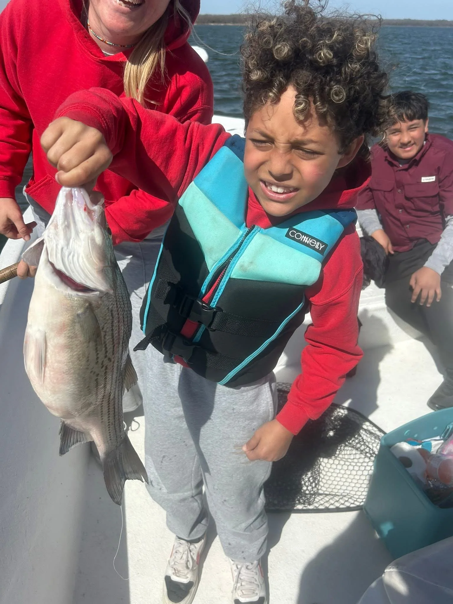 A person holding a fish on a boat with water in the background, and two children standing nearby, smiling.