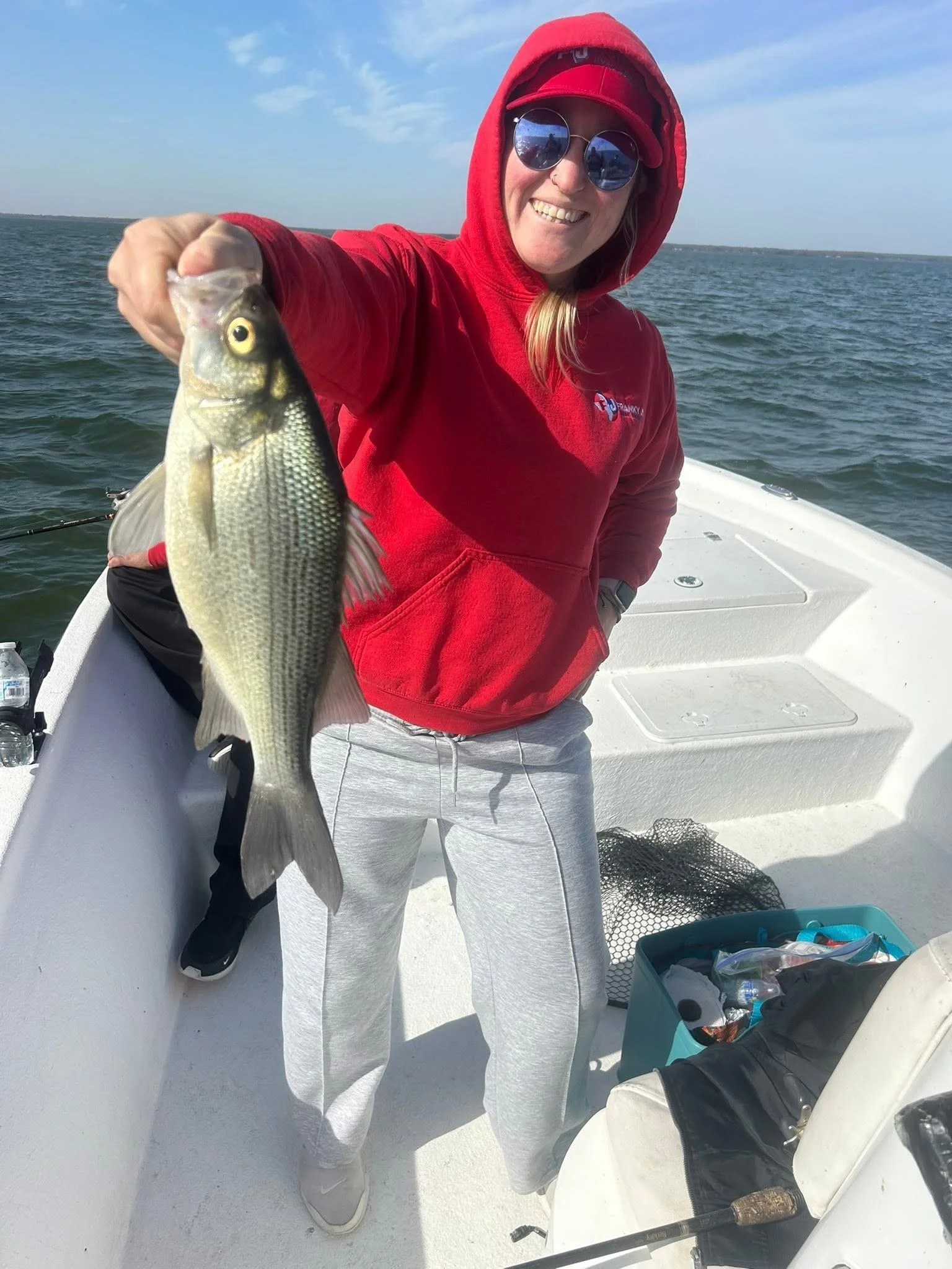 Woman in red hoodie holding a fish on a boat in a large body of water, smiling at the camera.