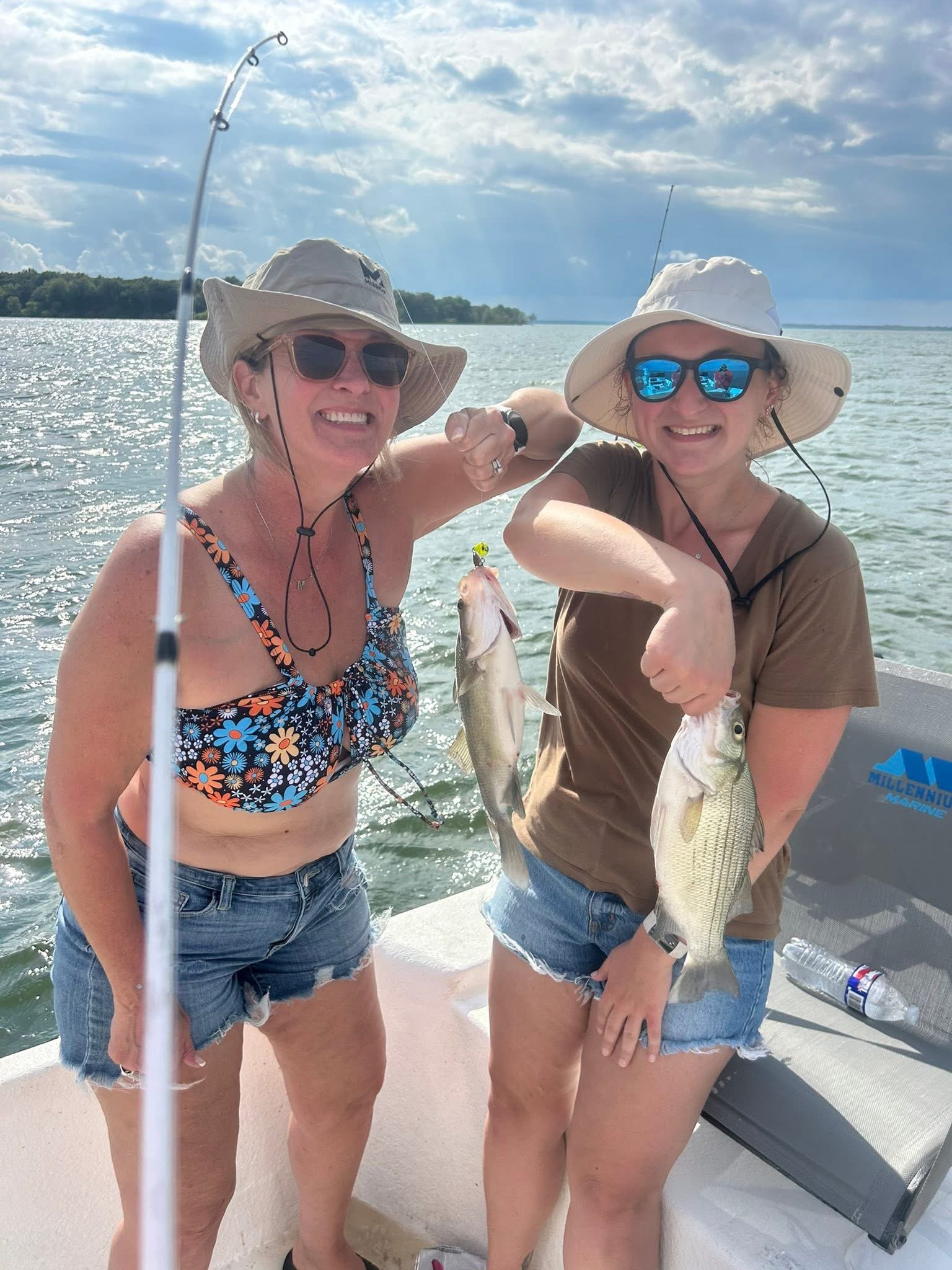 Two women in hats and sunglasses showing off two fish they caught on a boat with the water and sky in the background.