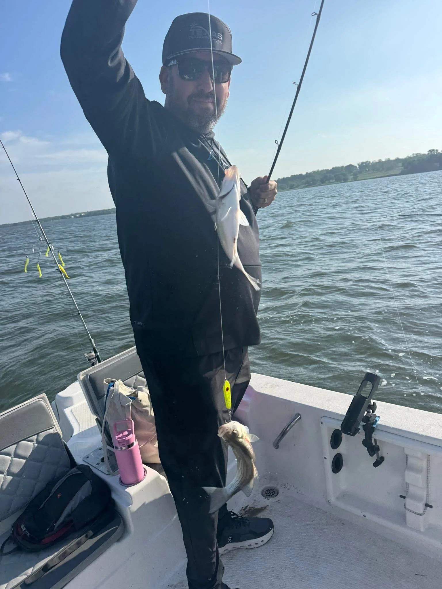 Man fishing on a boat holding a fish he caught, with fishing rods and gear visible in the boat, water and trees in the background.