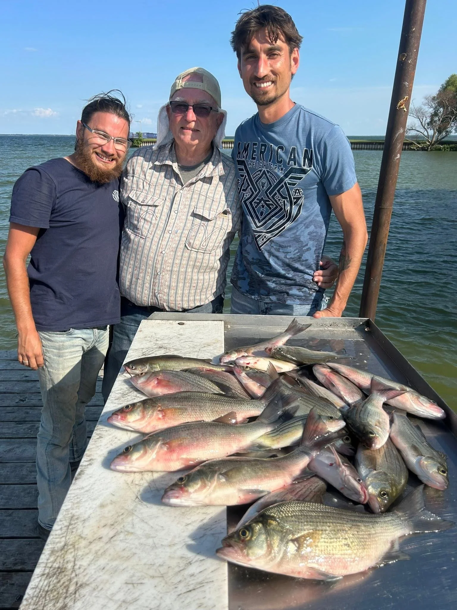 Three men standing behind a table with freshly caught fish, outdoors near a water body on a sunny day.