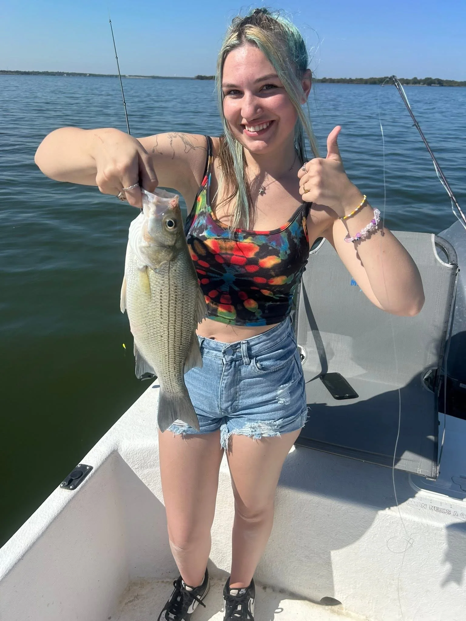 A young woman smiling and holding a fish on a boat in a body of water, giving a thumbs-up. She's wearing a colorful tank top, denim shorts, and black sneakers. The sky is clear and blue, with a distant shoreline visible.