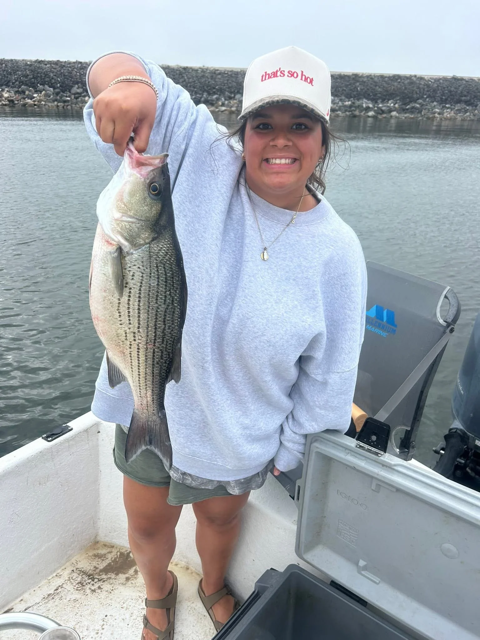Smiling woman holding a large fish on a boat near water with a rocky shoreline in the background.