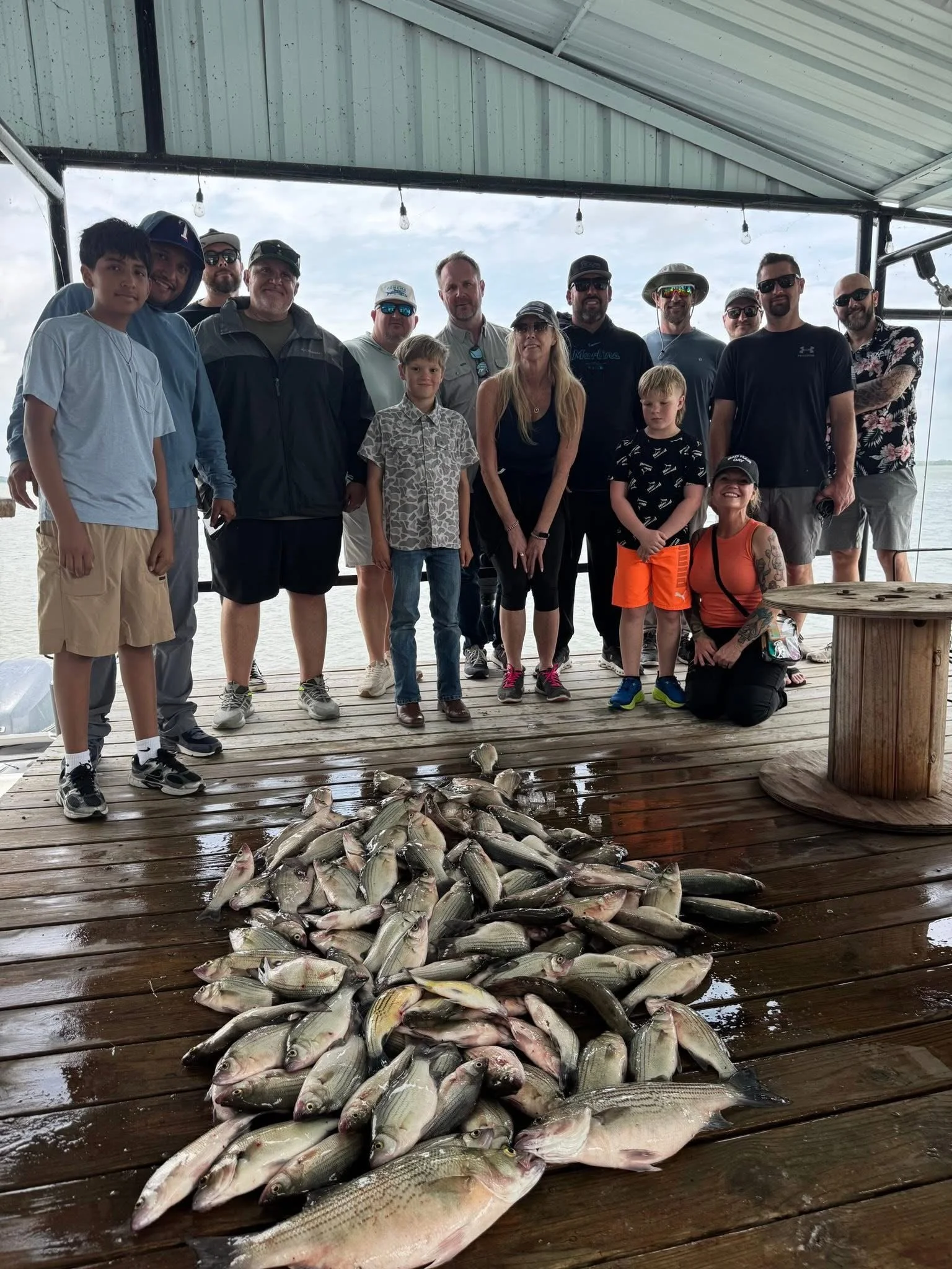 A group of people standing on a wooden dock in front of a body of water, with a pile of fish on the dock in front of them. The group includes adults and children, some wearing sunglasses and hats, and the sky is cloudy.