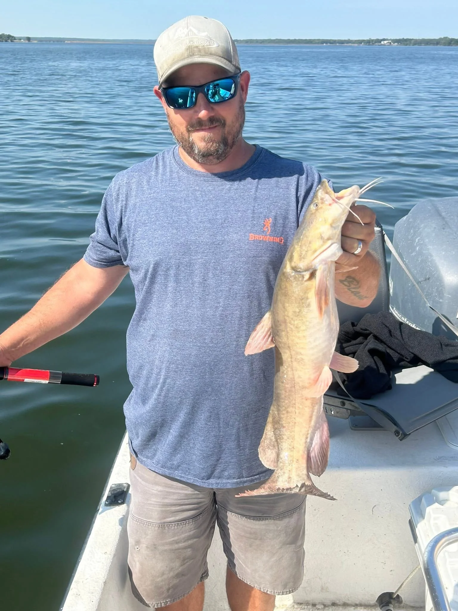 Man wearing sunglasses, cap, and gray t-shirt holding a large fish on a boat with water and distant shoreline in the background.