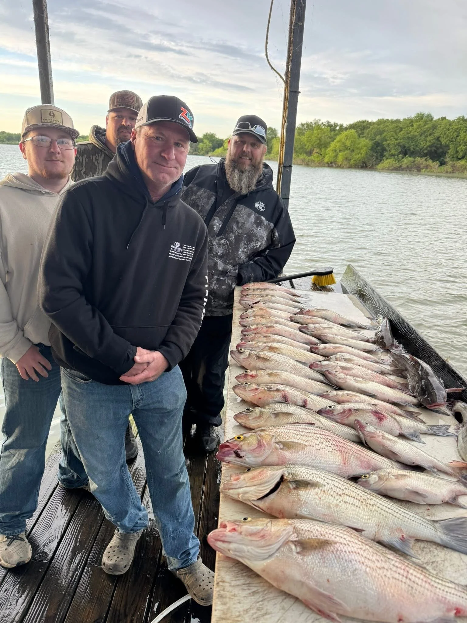 Four men standing on a dock beside a large catch of fish laid out on a table by the water, with trees in the background.