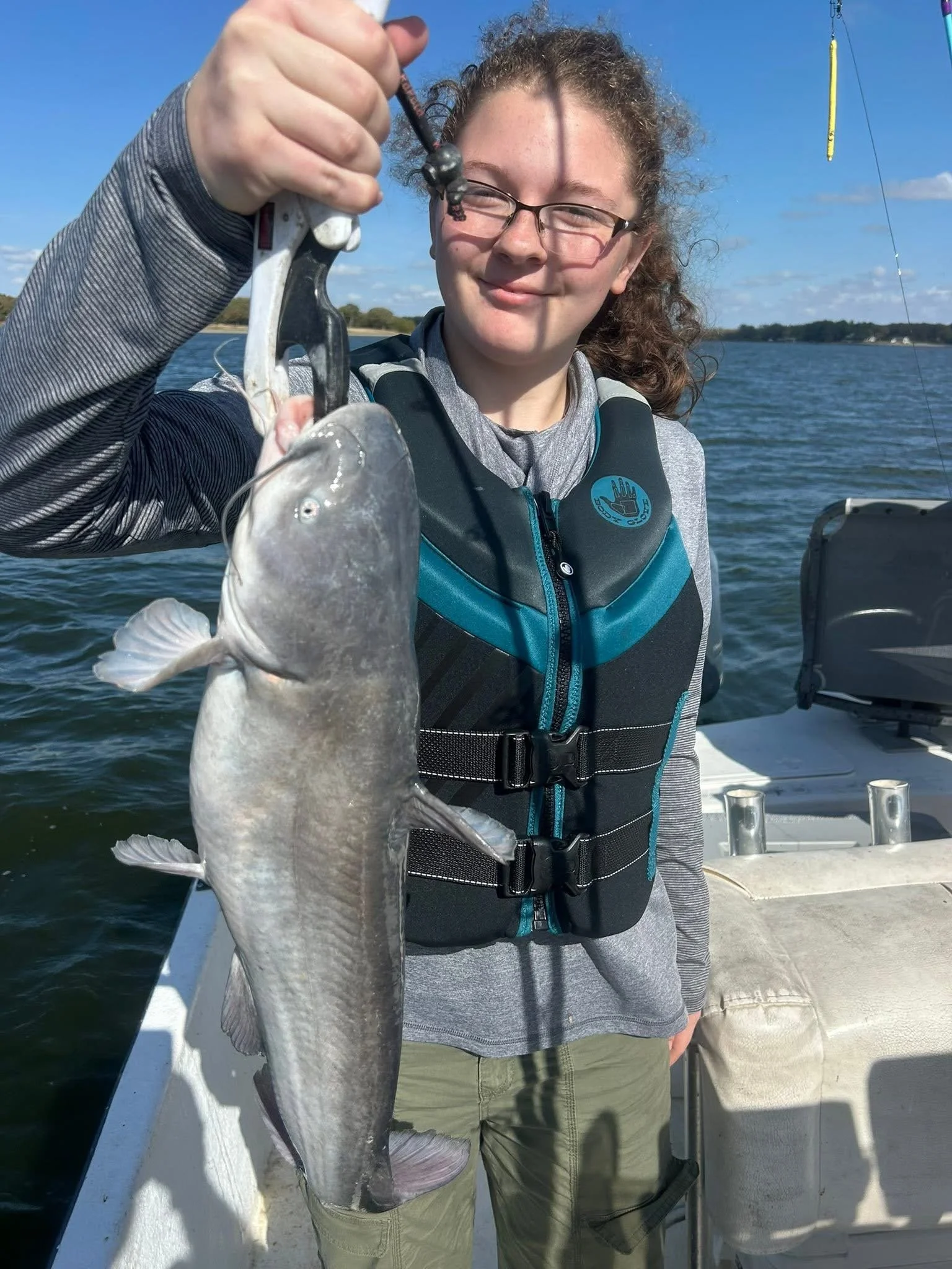 A woman holding a fish she caught while on a boat on a body of water, with a fishing rod visible in the background, during daytime with blue sky and some clouds.