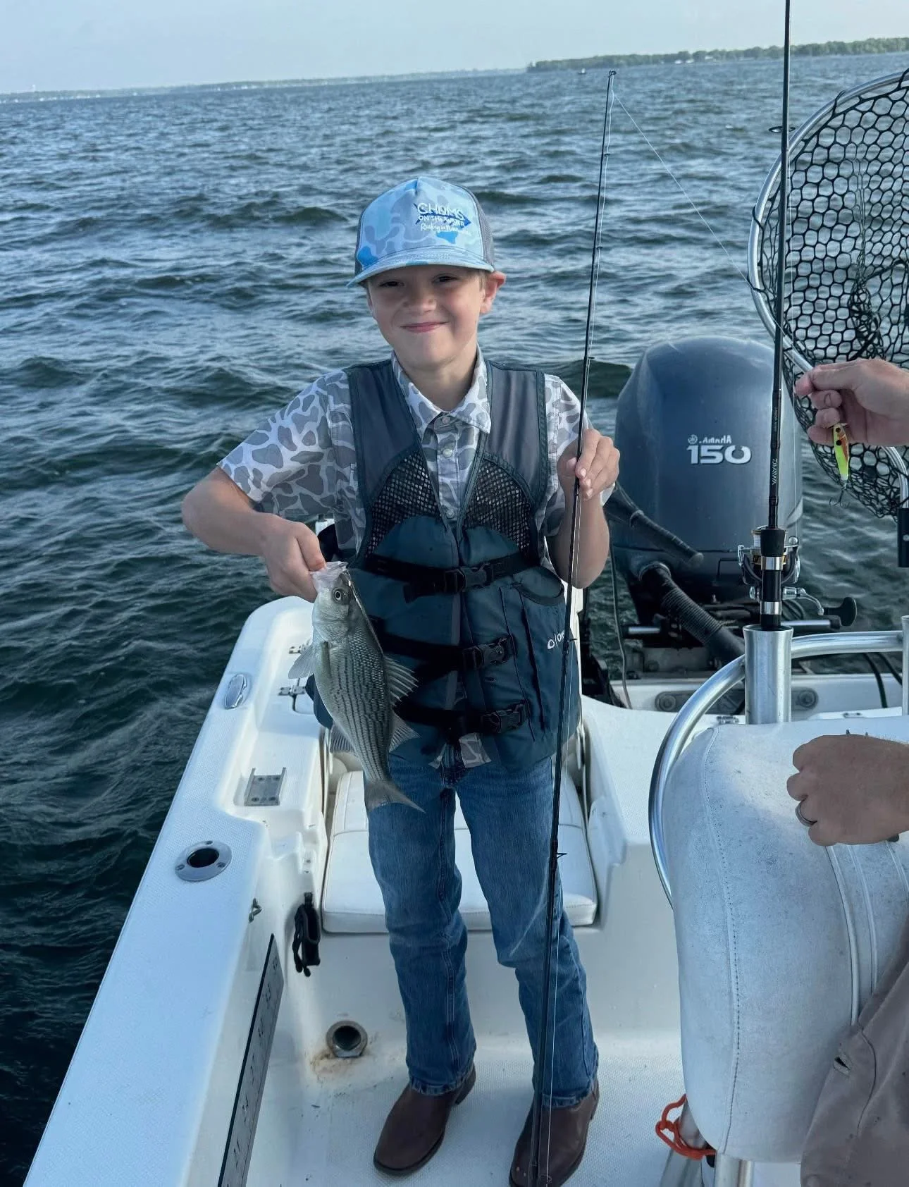 A young boy smiling and holding a fish on a boat, with open water and a distant shoreline in the background.