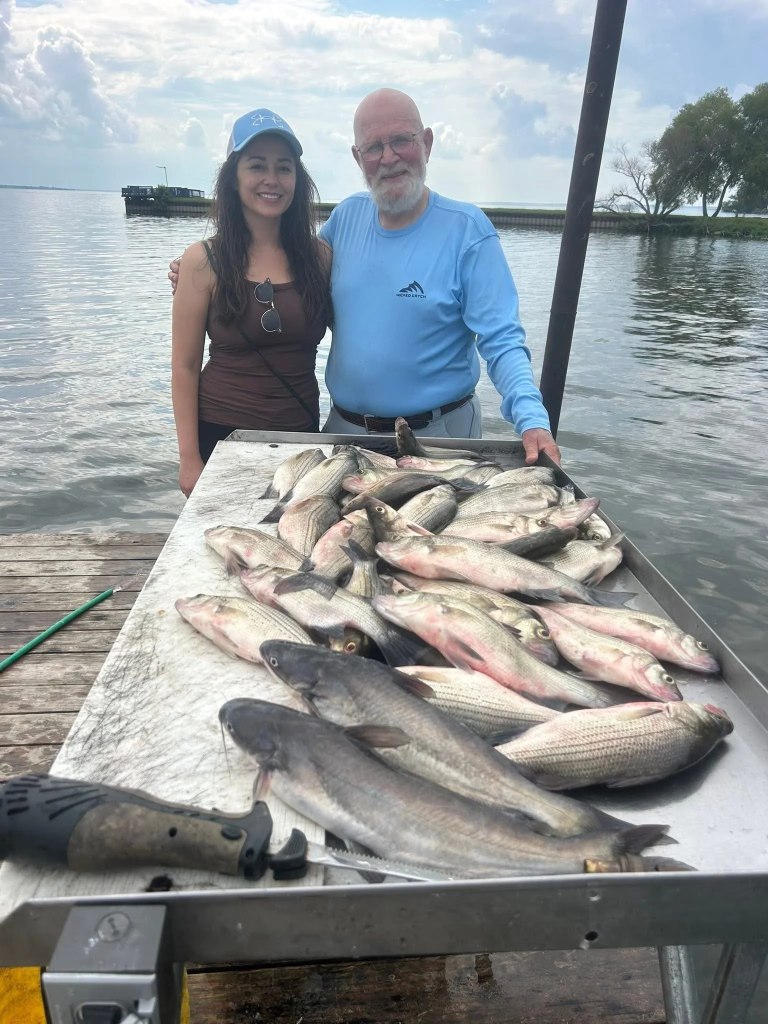 A young woman and an elderly man standing by a table full of numerous fish, next to a body of water, with a cloudy sky overhead.