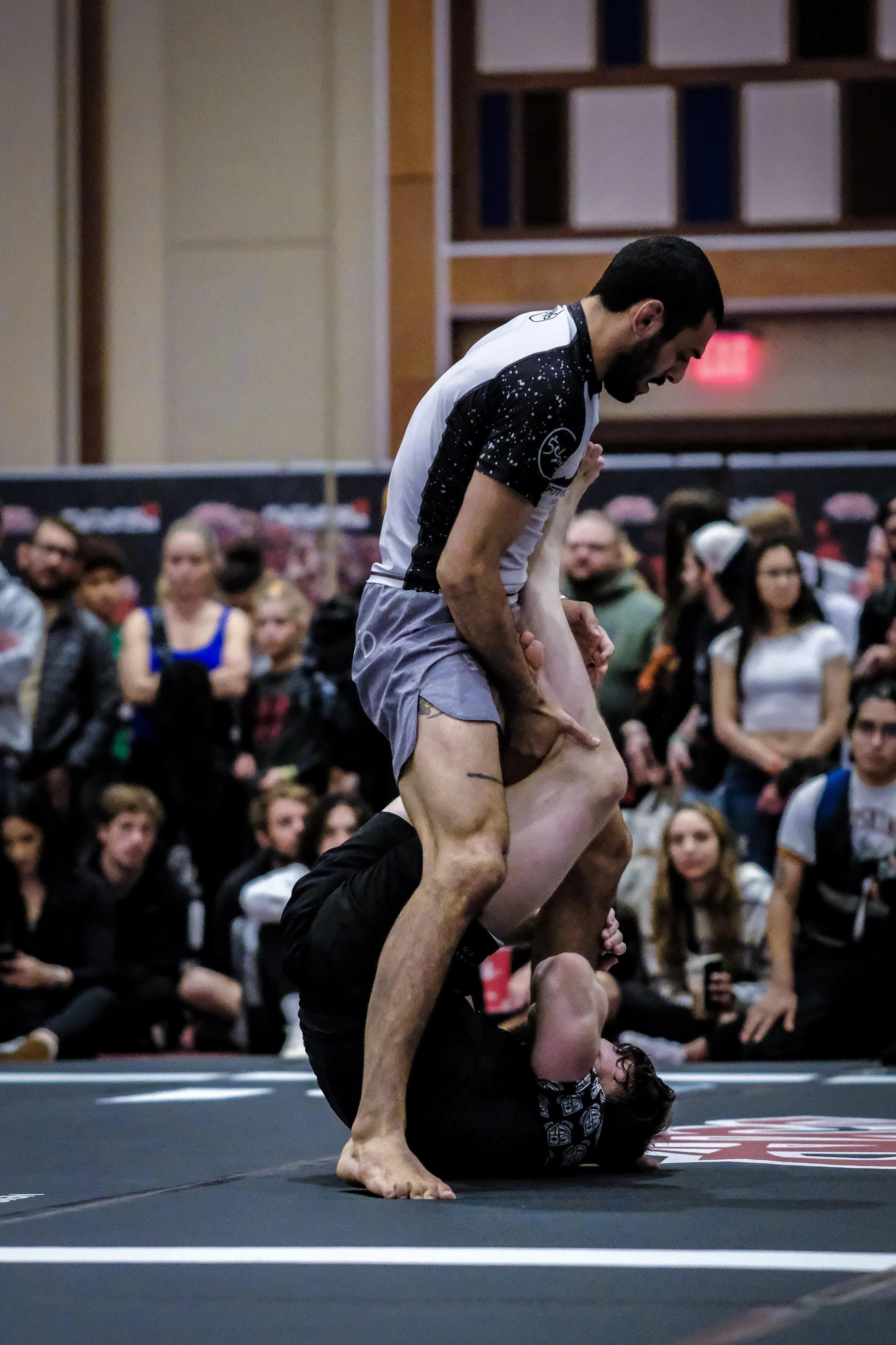 Two men practicing Brazilian Jiu-Jitsu on a black mat surrounded by spectators. One man is on top, gripping the other man around the waist, while the other man is on his back with legs over his opponent's shoulders.