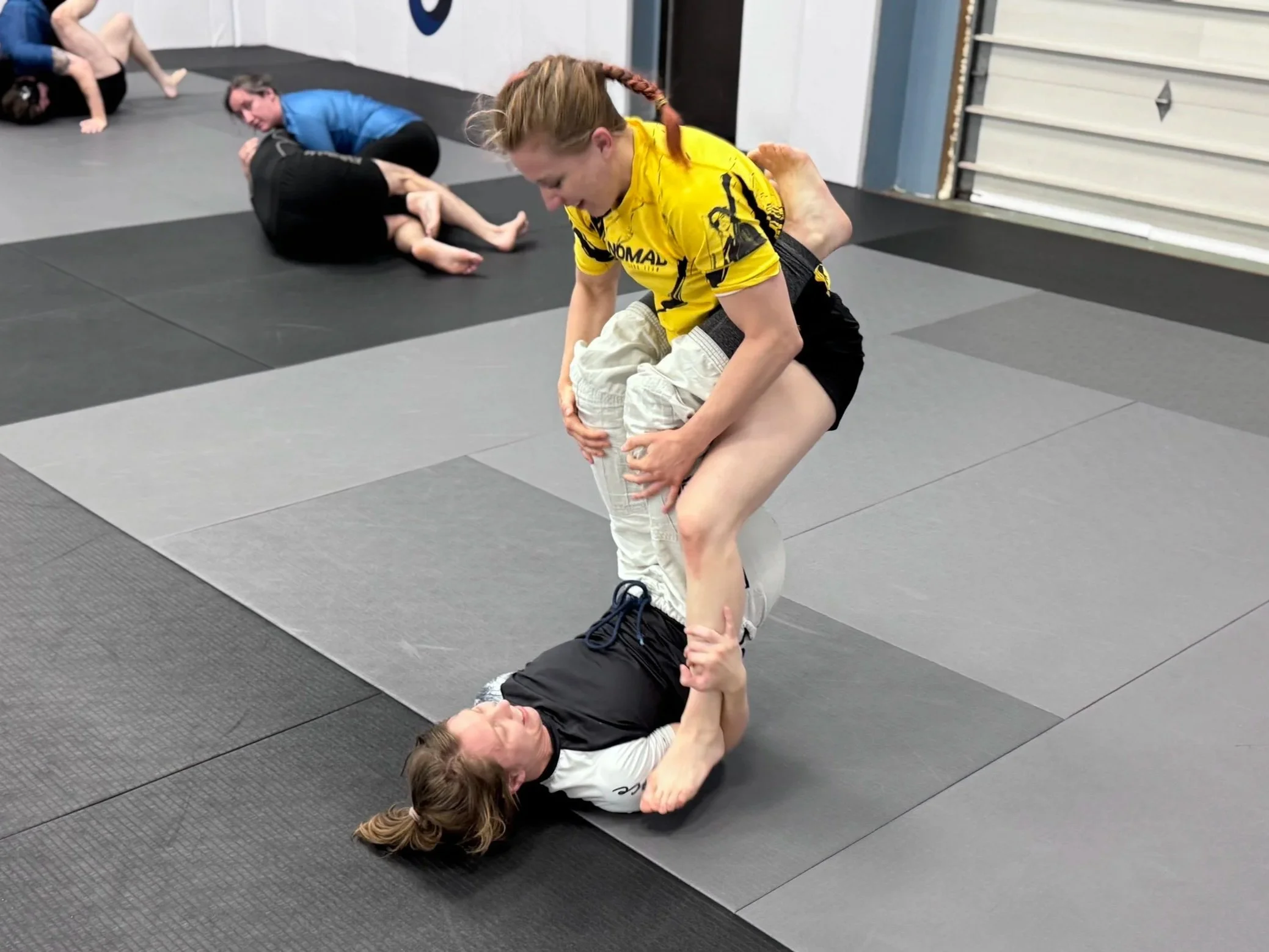 Two women practicing Brazilian Jiu-Jitsu on a black mat in a gym. One woman in a yellow rash guard and black shorts is standing with one leg lifted, preparing to pass the guard of the woman on the ground, who is in a black and white gi and is lying on her back. In the background, two other women are practicing GJJ techniques.