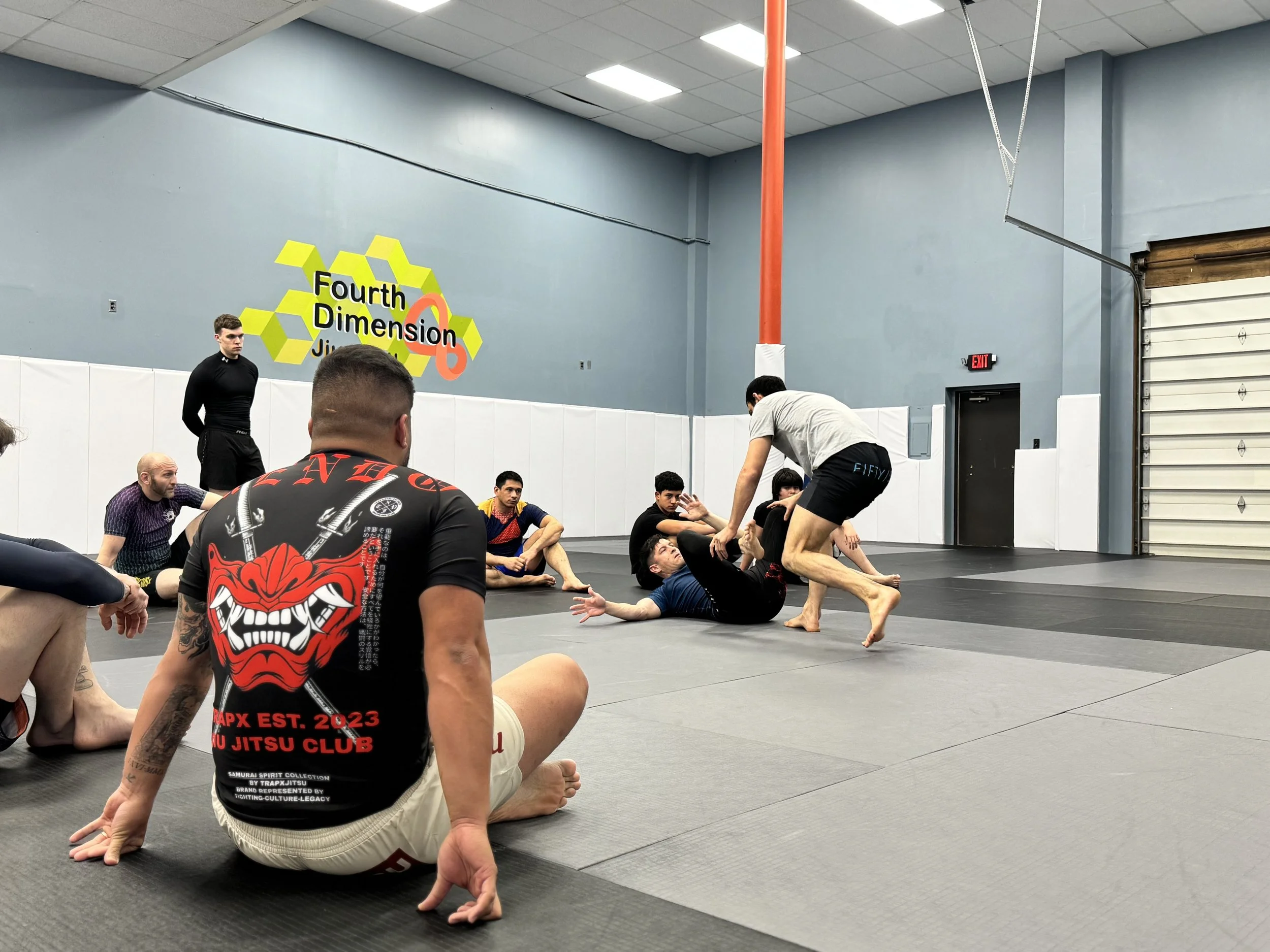 People practicing Brazilian Jiu-Jitsu on mats inside a martial arts gym. One person is sitting on the mat with others engaged in grappling techniques. The gym has light blue walls, a few posters and signs, and a garage-style door.
