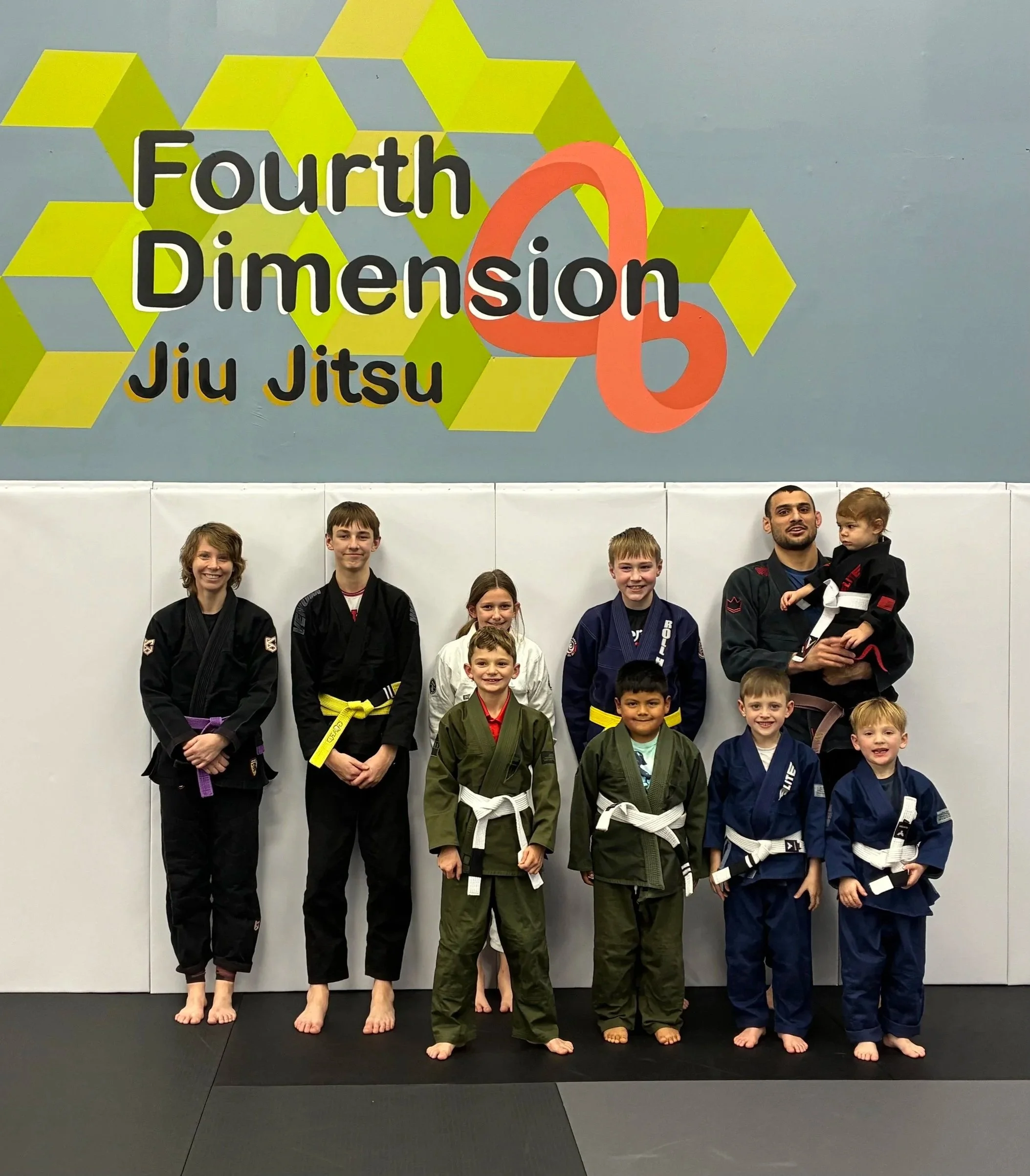 A group of children and their instructor pose for a photo in a martial arts class, standing on a black mat in front of a wall with a colorful "Fourth Dimension Jiu Jitsu" sign. The children wear gi uniforms with belts of various colors, and the instructor is in a black gi and holding a young child.