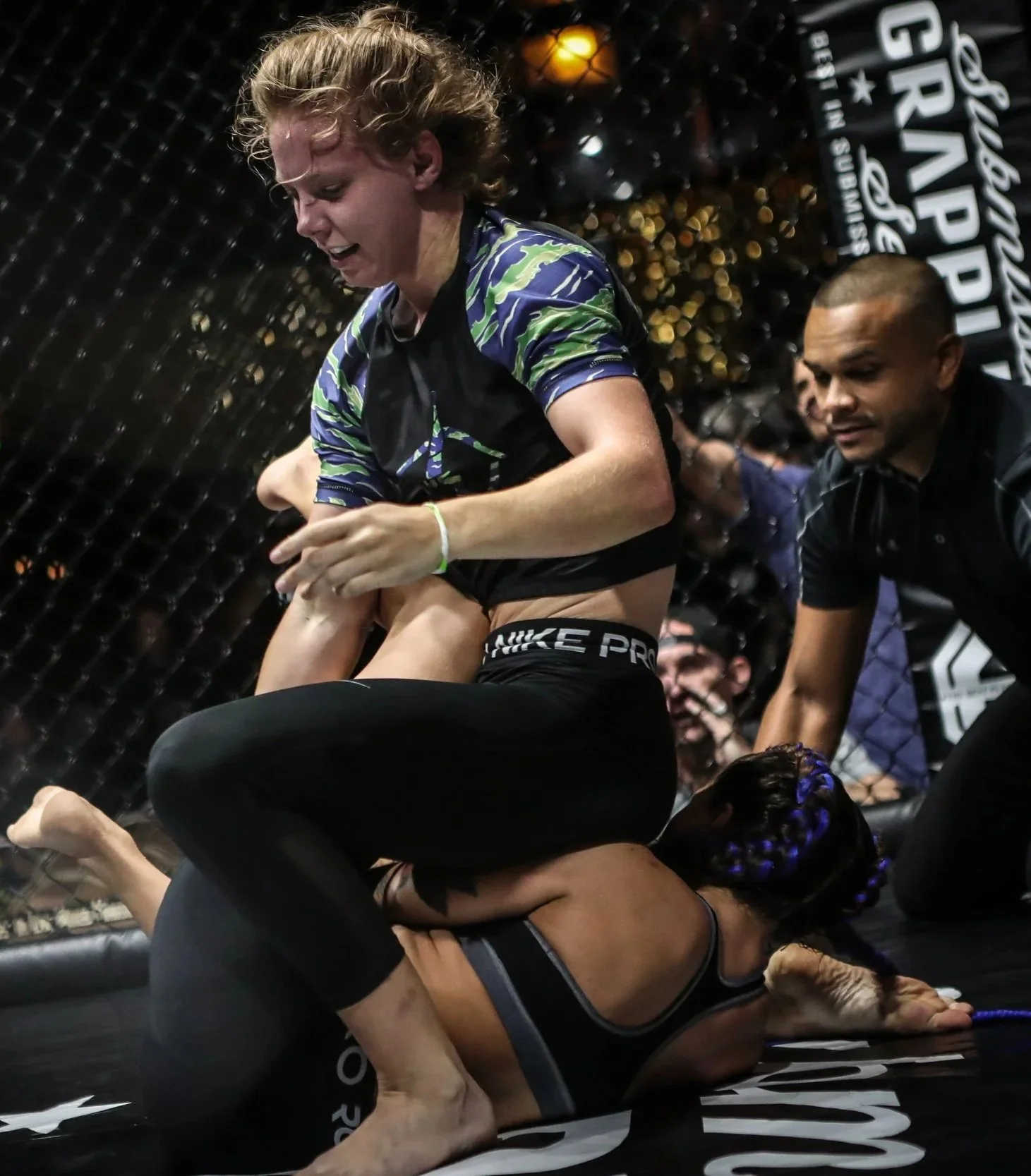 Mixed martial arts fighters in a grappling match inside a cage, with one fighter on top and another on the ground, surrounded by a referee and spectators.