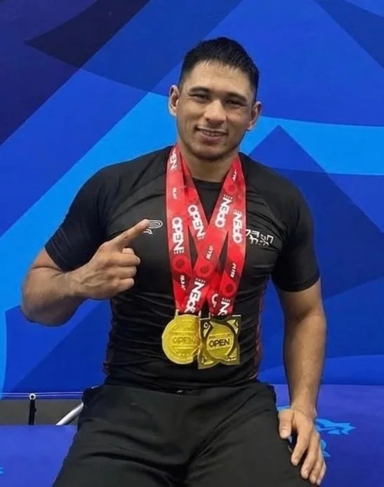 A young man with short dark hair smiling, wearing a black athletic shirt and black shorts, sitting in front of a blue geometric background, pointing to gold medals around his neck.