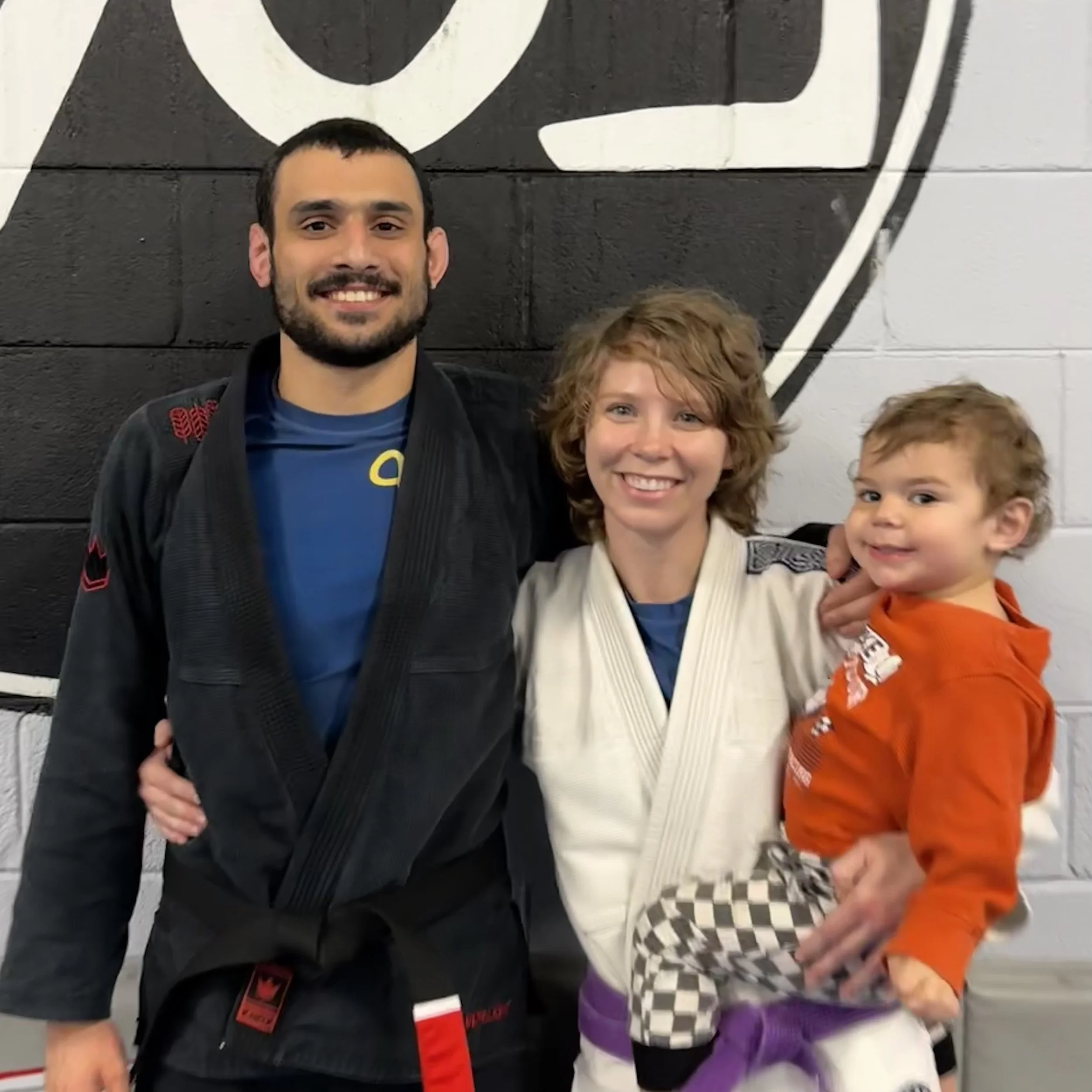 Close-up of a smiling young man with short dark hair and a beard, standing with two children in front of a black and white wall with artwork. One child has curly light brown hair and is wearing a white martial arts uniform with a black belt, and the other child has short light brown hair and is wearing a bright orange hoodie.