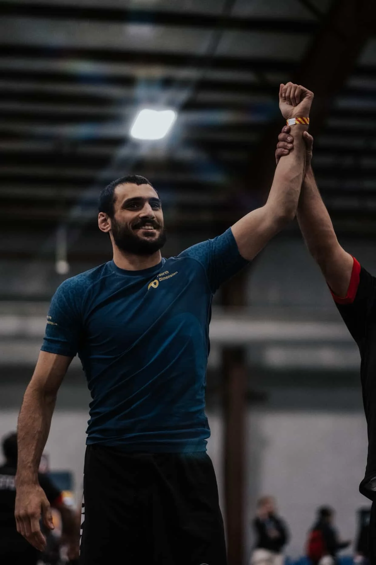 A man with a beard in a blue athletic shirt and black shorts smiling as he raises his arm in victory, holding hands with another person during an indoor sports event.