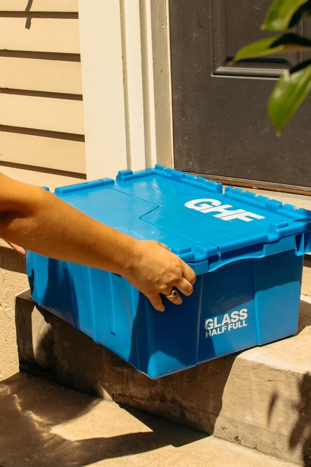 A person placing a blue plastic container labeled "GHF" with a dished lid outside a house. The container has markings "GLASS HALF FULL" on the side, and is positioned on a concrete step in front of a door.