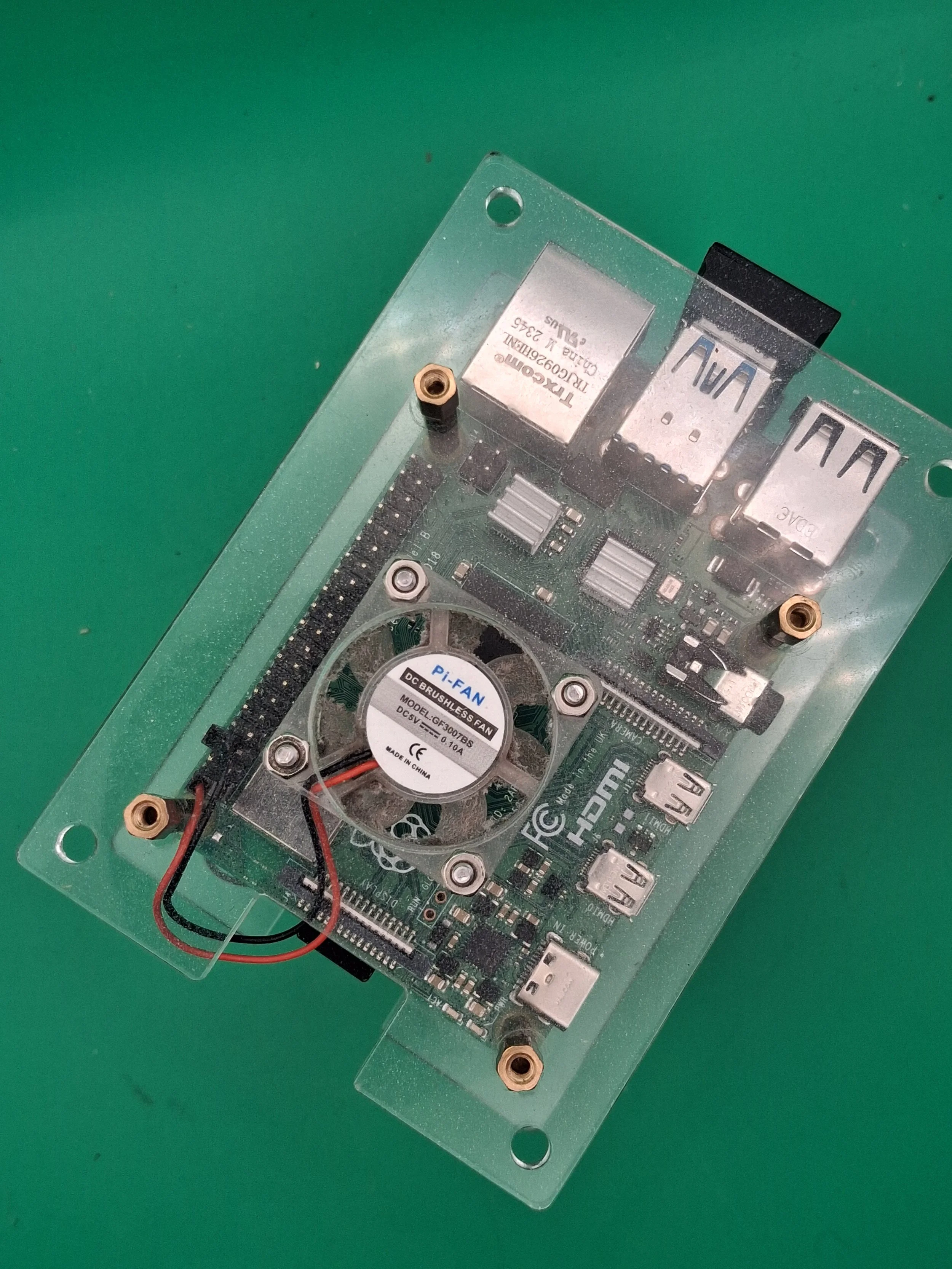 A close-up view of a green printed circuit board with various electronic components, including a small cooling fan labeled 'Pi-FAN' and two USB ports, mounted within a transparent protective case with brass standoffs.