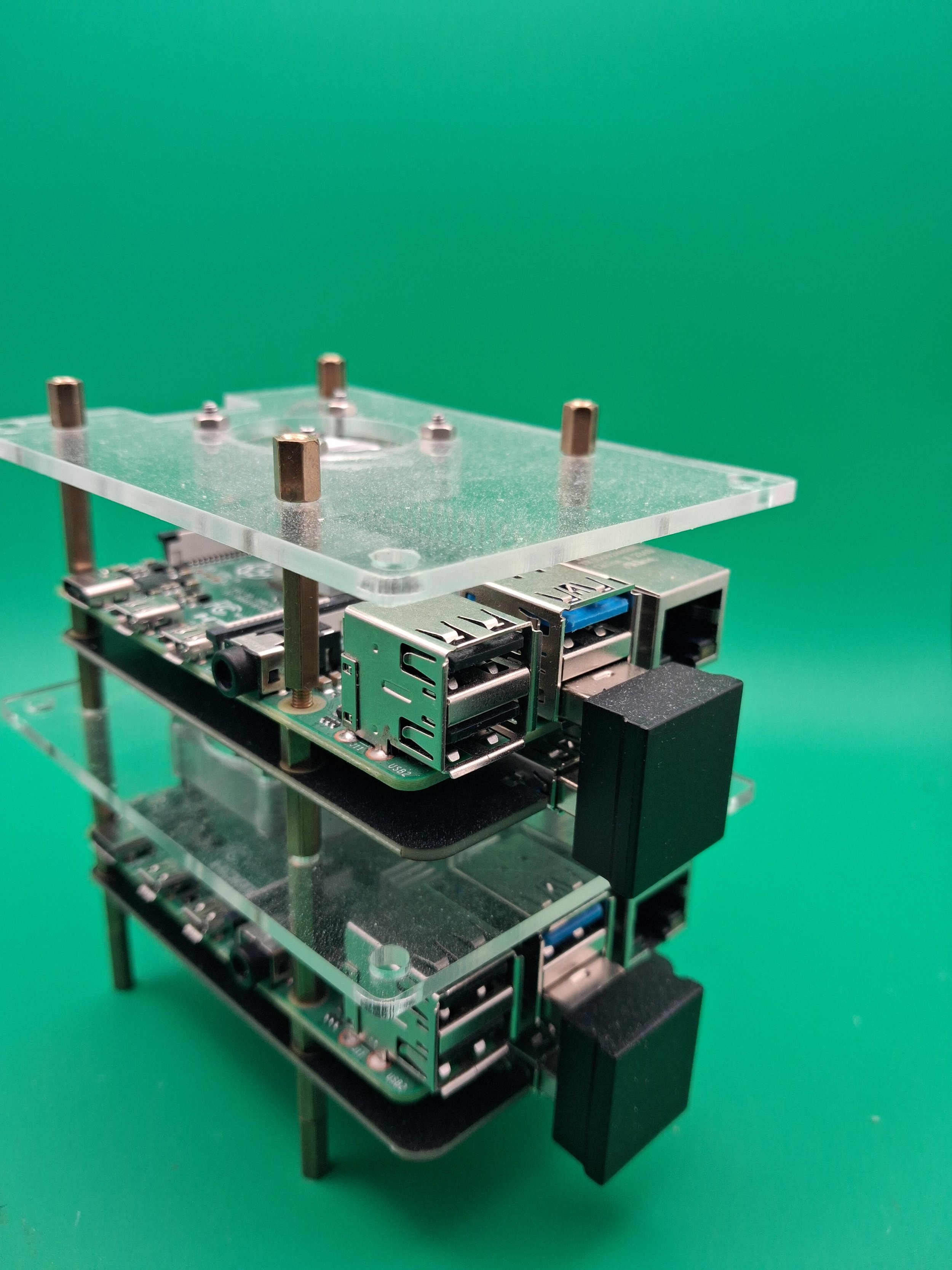 A Raspberry Pi computer mounted on a small stand with a clear acrylic top, showing metal standoffs, and a black heatsink attached to the side, set against a green background.