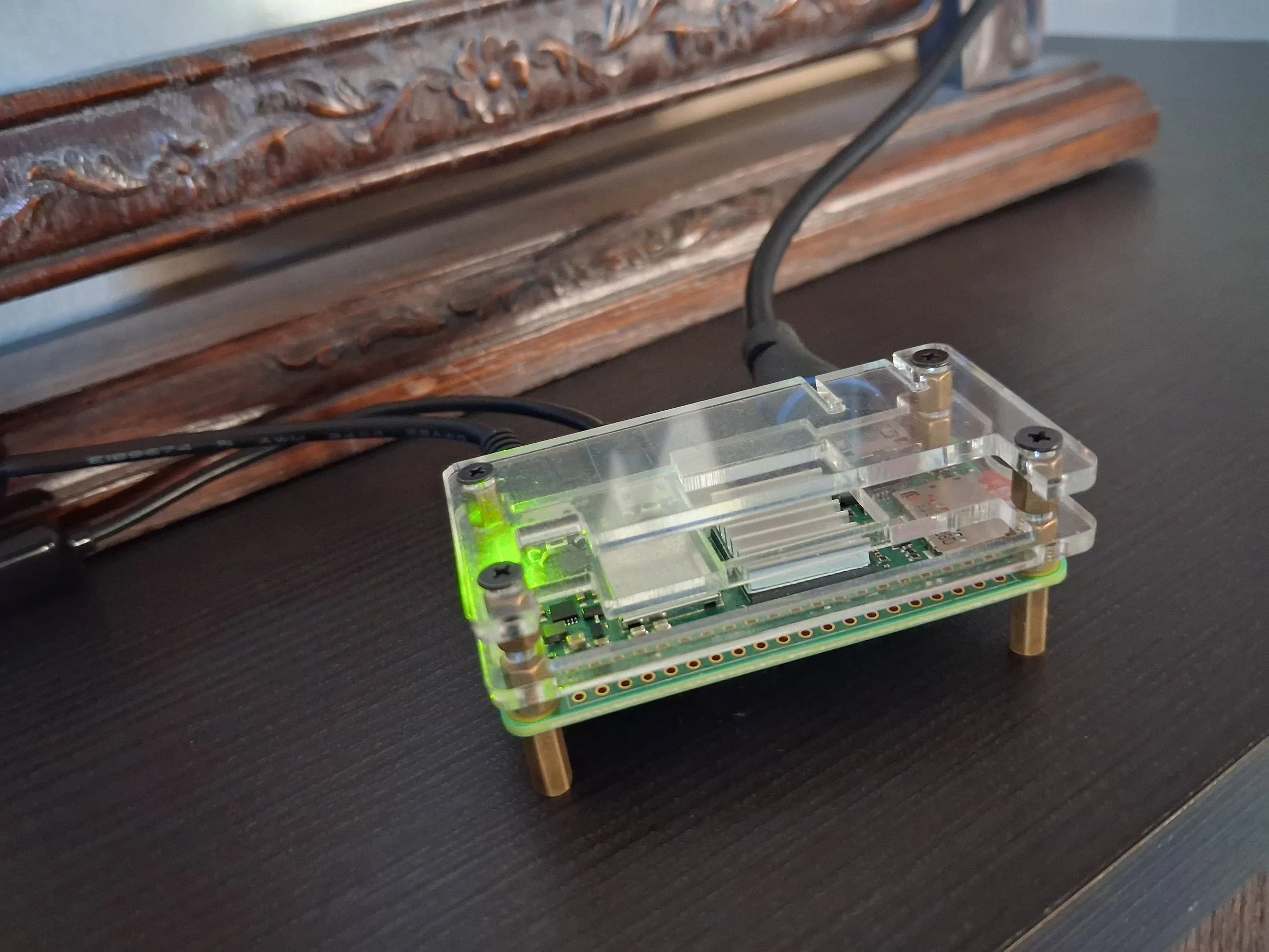 A clear plastic electronic device with green LED lights, connected by a black cable, sitting on a black surface near a carved wooden decorative object.