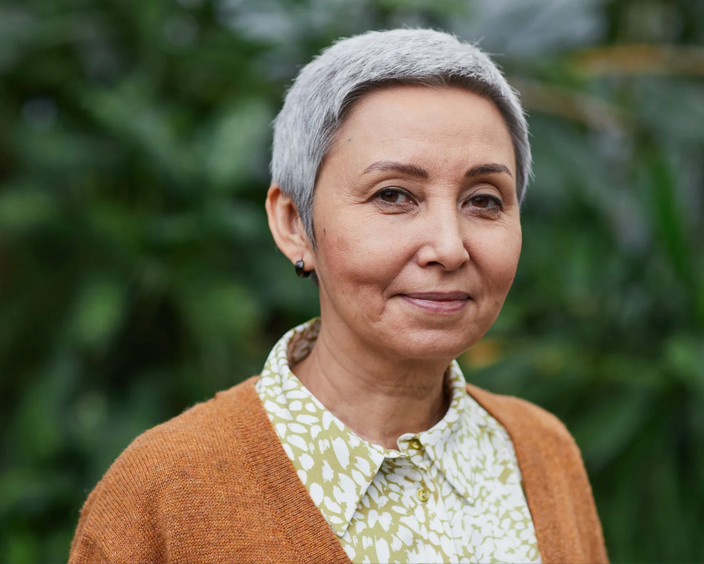 An older woman with short gray hair, wearing a patterned blouse and a brown cardigan, standing outdoors with green foliage in the background.
