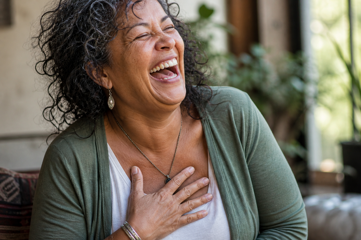 A woman with curly black hair, wearing earrings, a necklace, and a green cardigan, laughing heartily with her hand on her chest in a bright, cozy room with plants in the background.