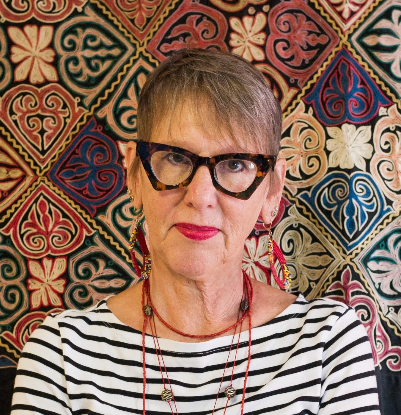 A woman with short brown hair, wearing tortoiseshell glasses, red lipstick, and colorful jewelry, standing in front of a vibrant, patterned textile background.