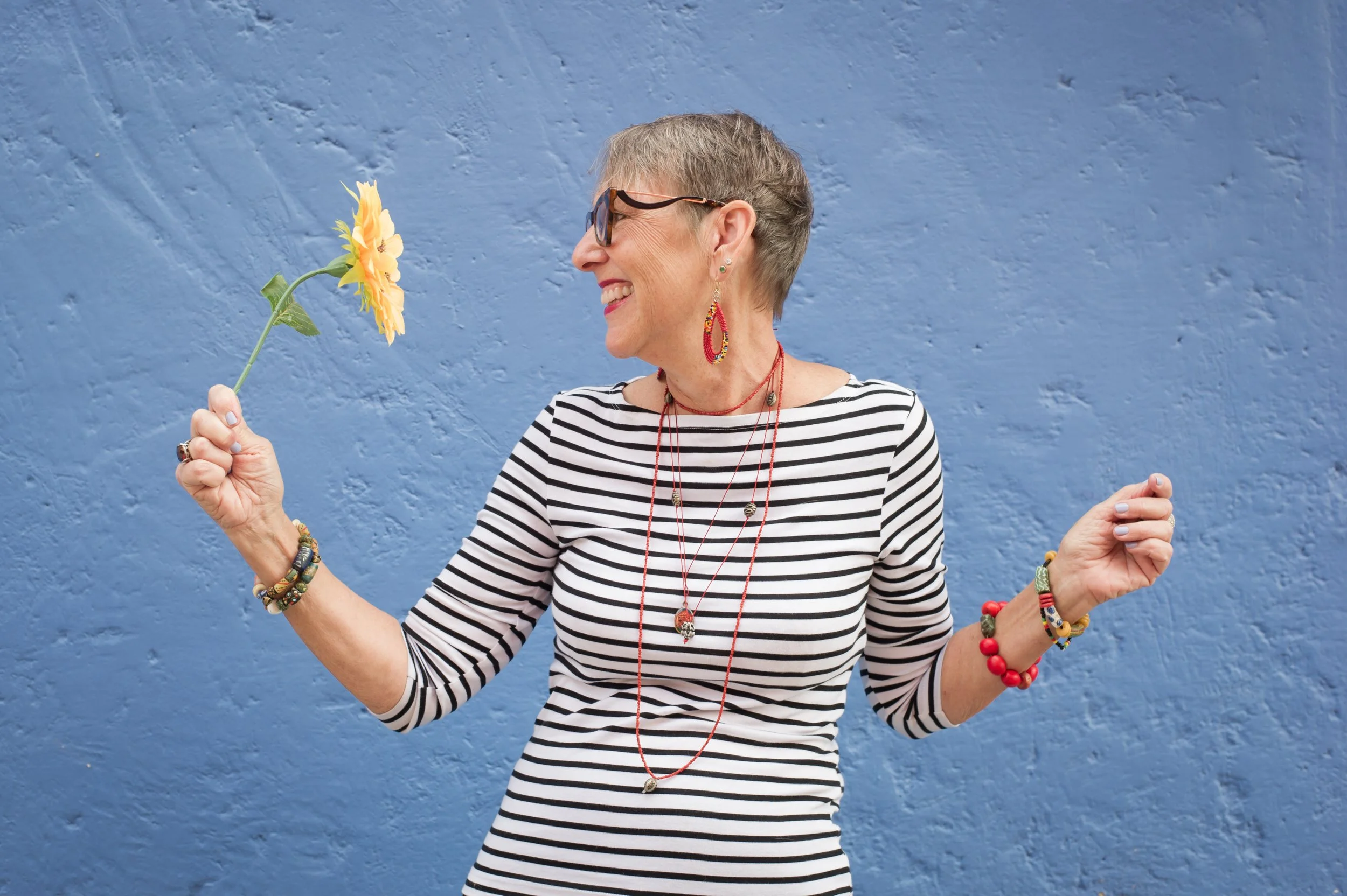 A woman with short gray hair, glasses, and colorful jewelry is smiling while holding a yellow sunflower in front of her face. She is wearing a black and white striped shirt and standing in front of a textured blue wall.