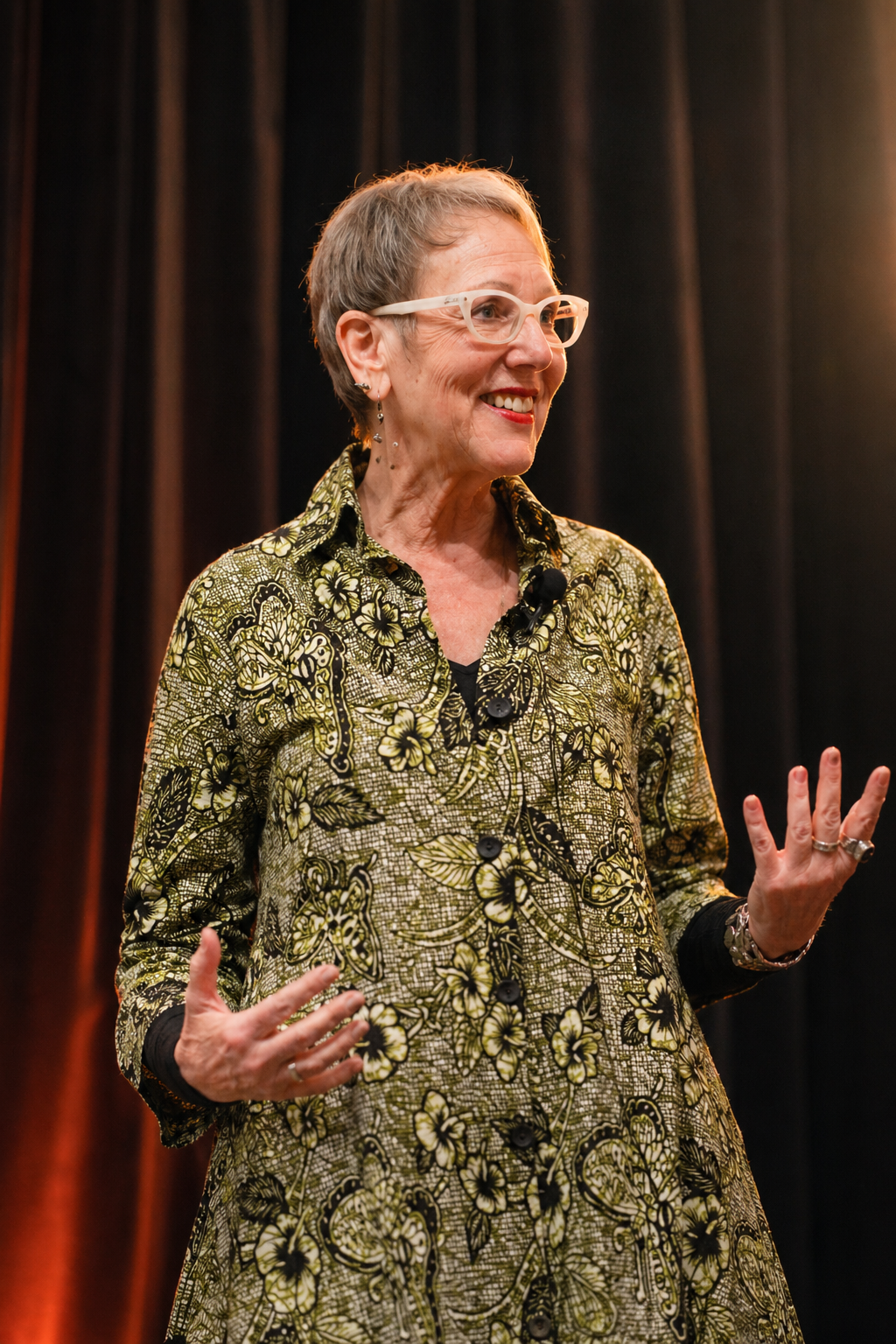 A smiling woman with short hair and glasses, wearing a patterned dress, speaking on a stage with dark curtains in the background.