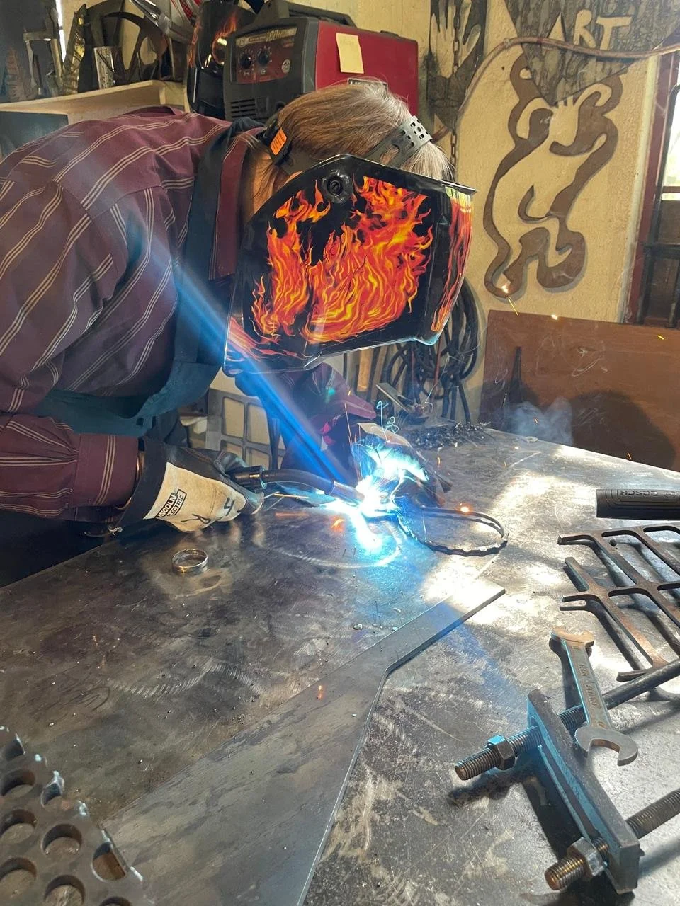 A person welding metal at a workbench, wearing a protective helmet with a flame design, gloves, and safety gear in a workshop.