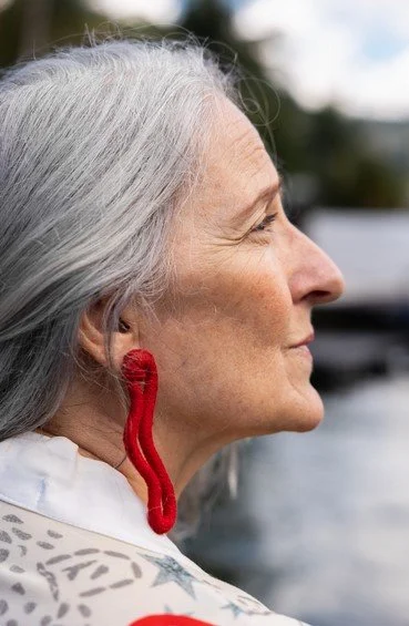 Profile of elderly woman with long gray hair and red decorative earrings, outdoors near water.