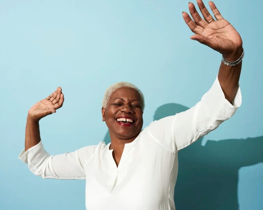 An older woman with short, platinum blonde hair, wearing a white long-sleeve shirt, smiling and raising her arms in a joyful manner against a blue background.