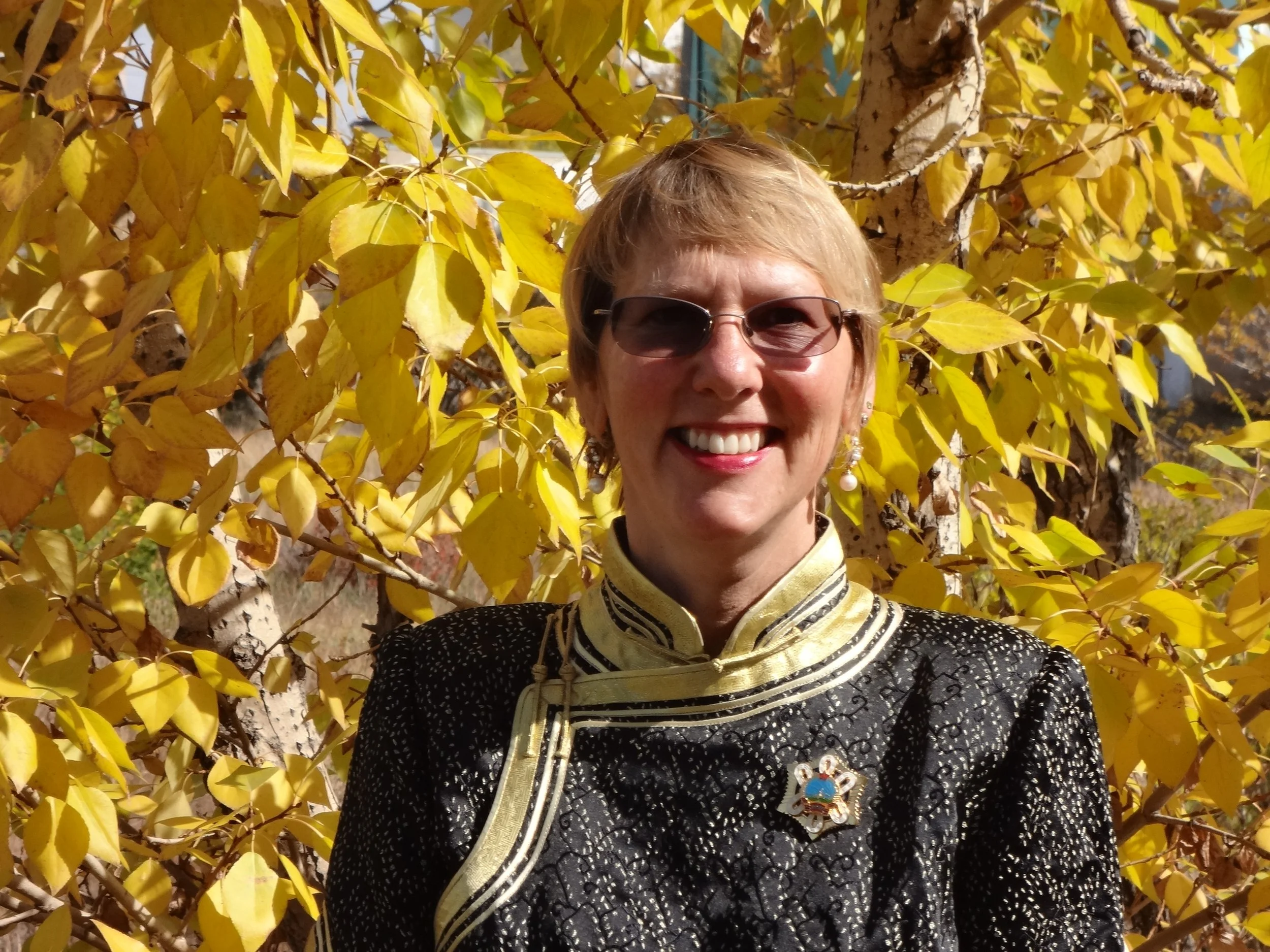 A smiling woman with short blonde hair wearing sunglasses, earrings, a brooch, and a decorative scarf, standing outdoors in front of a tree with yellow leaves.