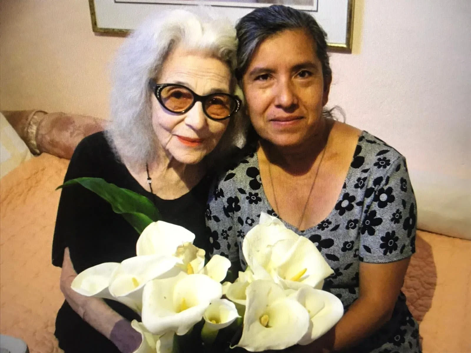Two women sitting on a sofa, holding a bouquet of white flowers, smiling at the camera; one woman has gray hair and glasses, the other has dark hair and is wearing a gray floral shirt.
