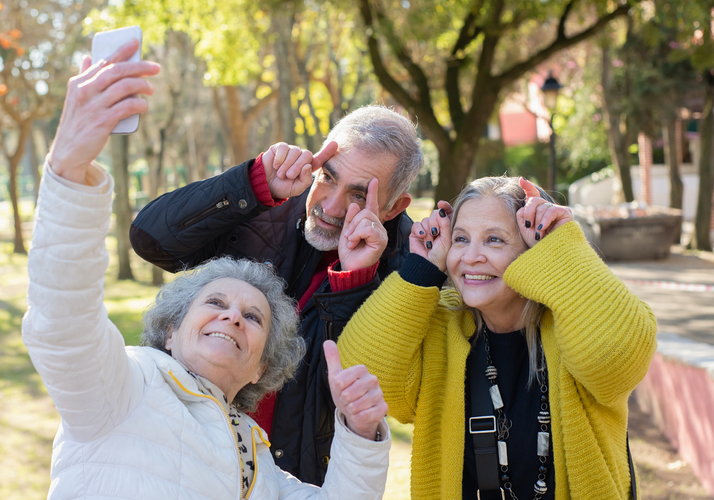 Three seniors taking a selfie outdoors in a park, smiling and making playful gestures, with trees and a walking path in the background.