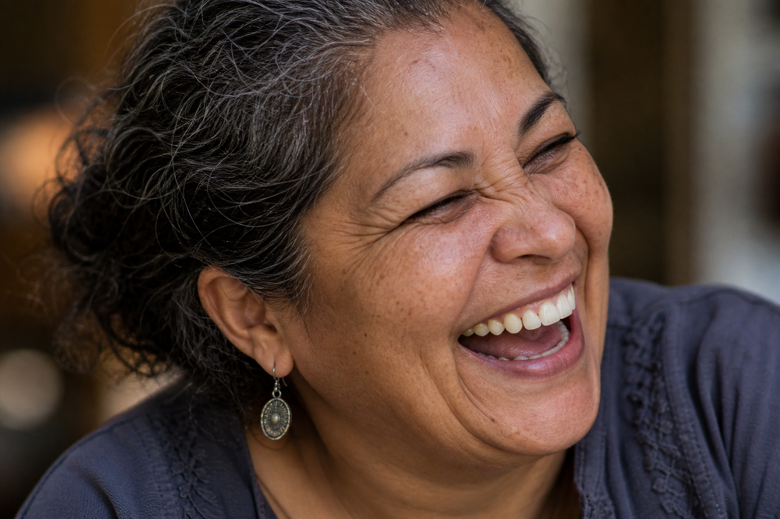 Close-up of a woman with salt-and-pepper hair smiling and laughing, wearing earrings and a dark shirt.