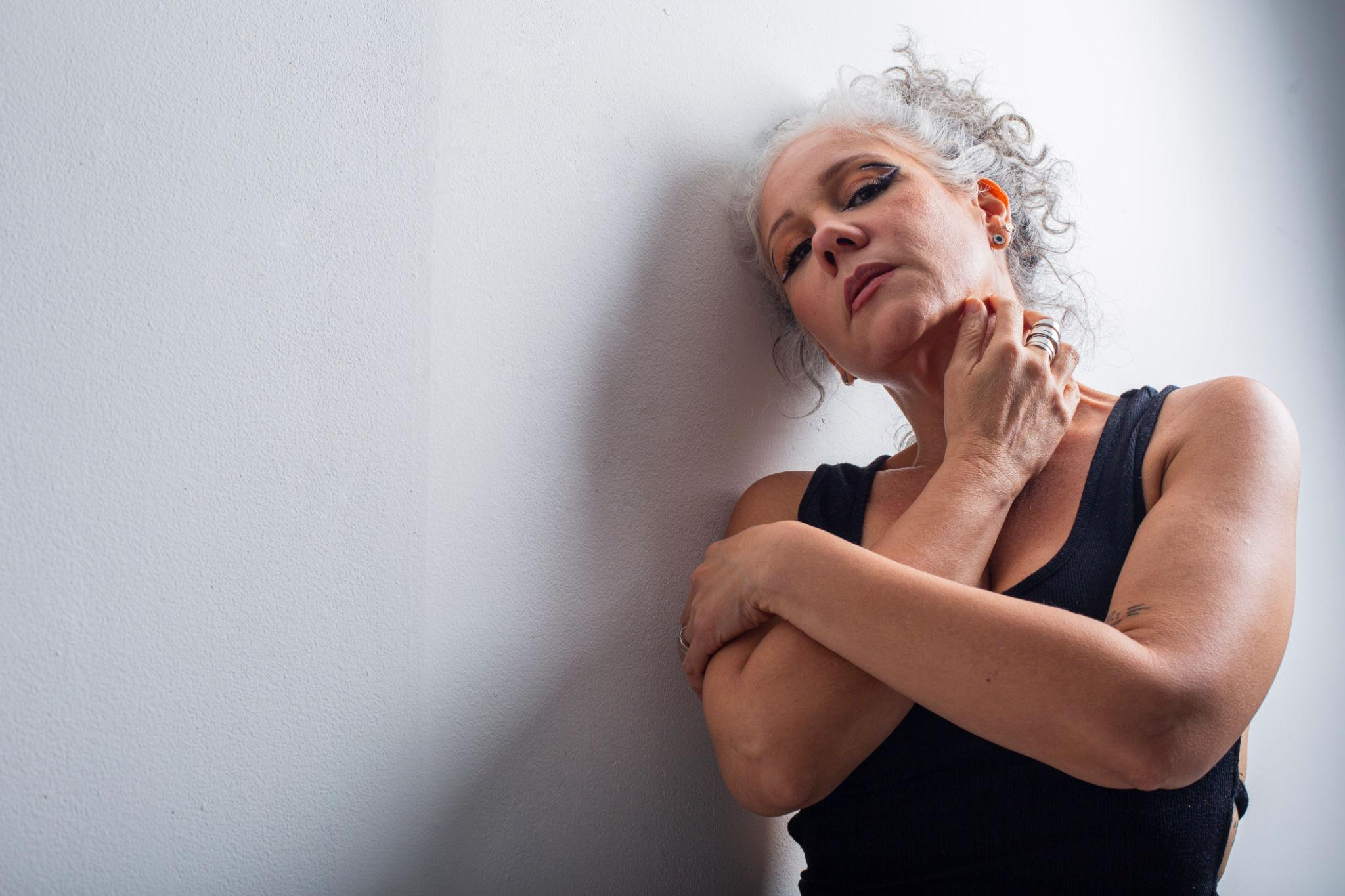 A woman with curly gray hair and dark eye makeup stands against a white wall, with her hand on her chin and her arms crossed. She wears a black sleeveless top and multiple rings.
