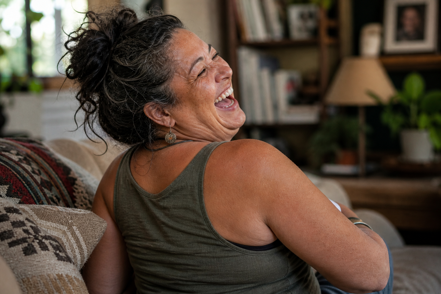 A woman with curly gray hair laughing and smiling while sitting on a sofa in a cozy living room.