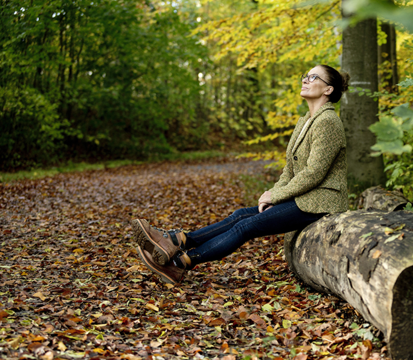 A woman with glasses, wearing a green patterned jacket and jeans, sitting on a large fallen tree trunk in a forest with autumn foliage, relaxing and enjoying the sunlight.