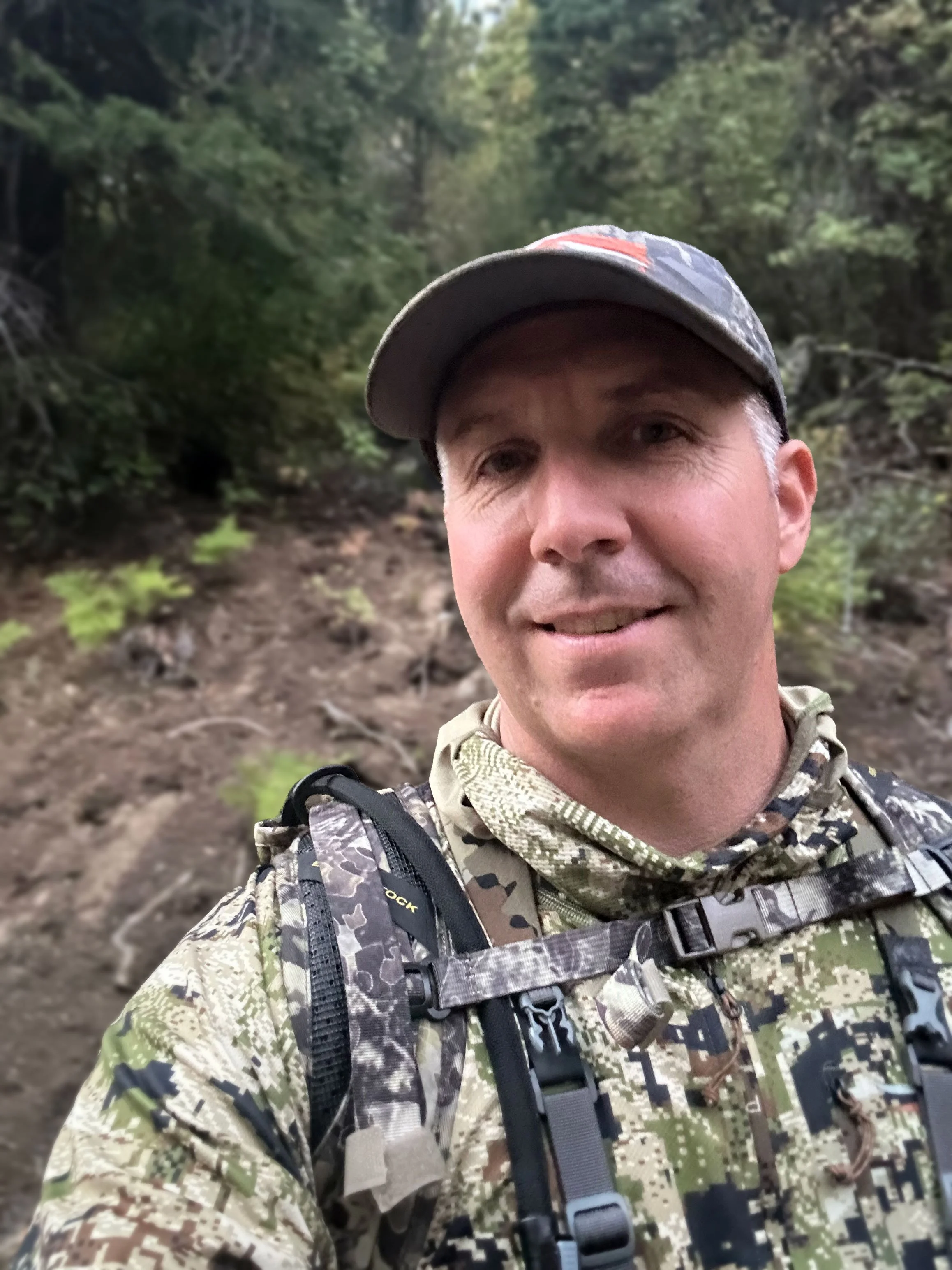 A man wearing camouflage military gear, including a camouflage hat, backpack, and tactical equipment, taking a selfie in a forested area with green trees and a dirt trail.