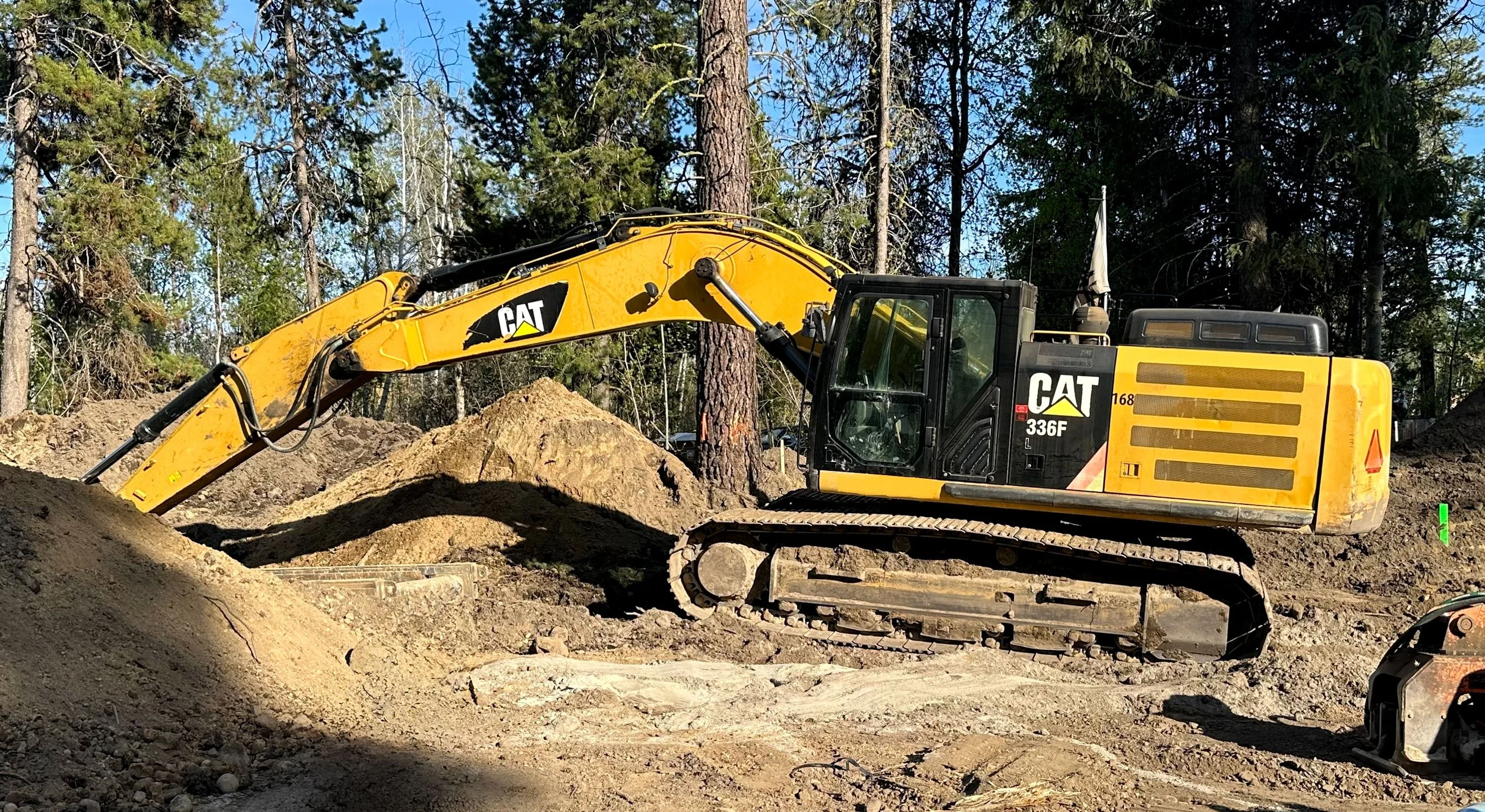 Yellow Caterpillar excavator with black tracks working on a dirt site surrounded by trees.