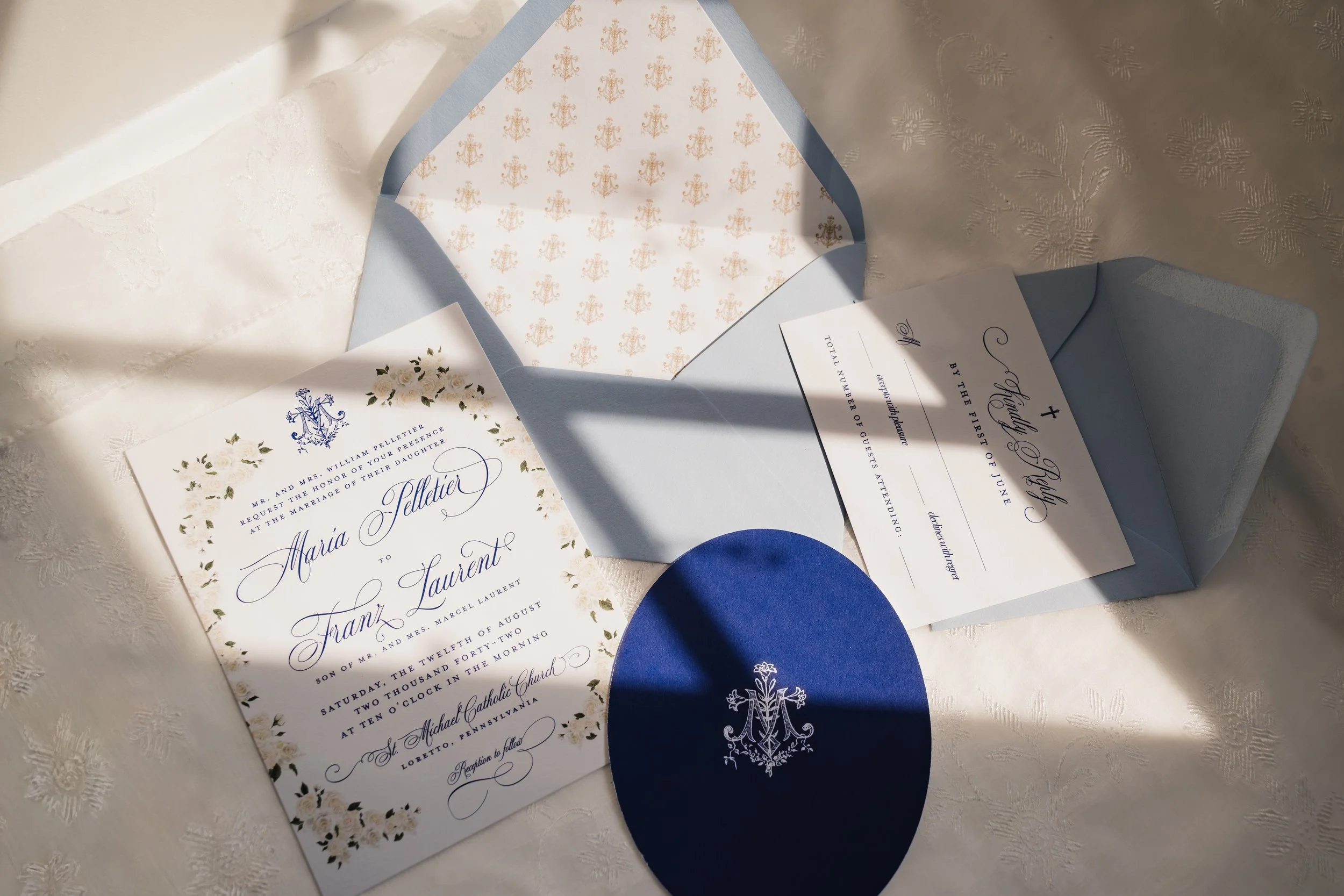 A blue, white, and cream Catholic wedding invitation suite is pictured with a dramatic shadow from natural lighting enhancing the visual effect.