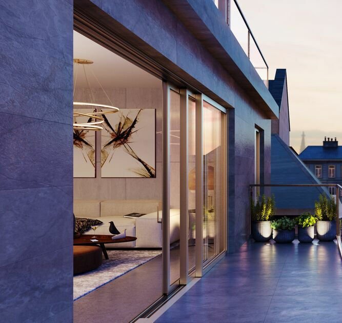 A modern balcony with glass sliding doors leading to a living room, decorated with potted plants and overlooking neighboring buildings at sunset.