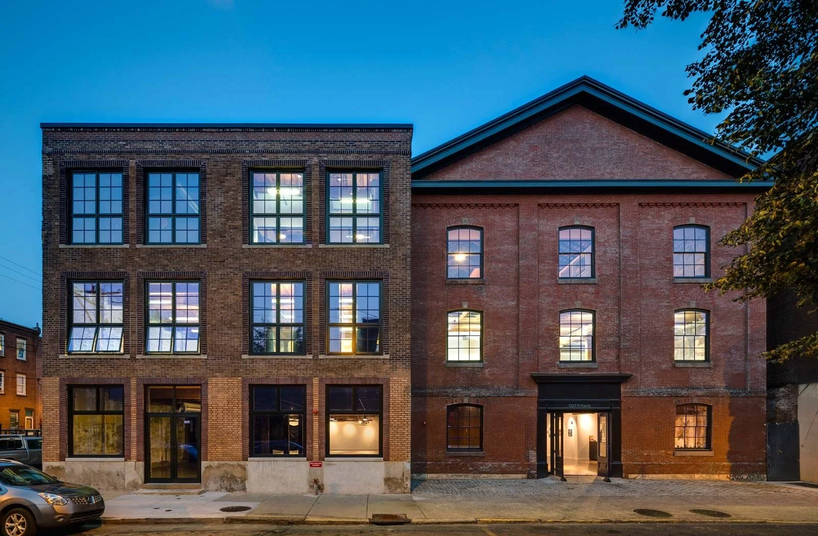 Two adjacent brick buildings at dusk, one with modern black-framed windows and the other with traditional arched windows, with interior lights on.