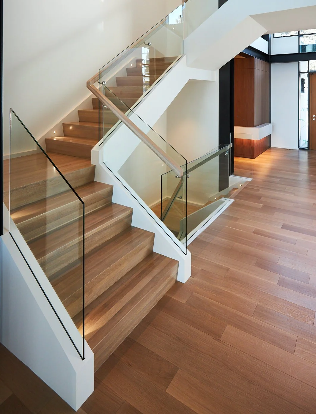 Modern interior with wooden stairs, glass railings, and hardwood flooring in a well-lit entryway.